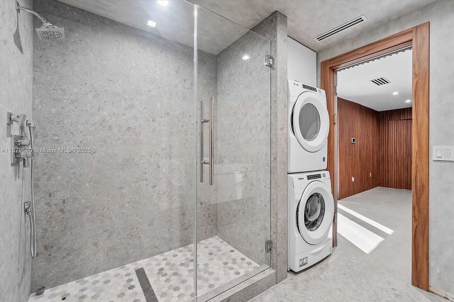 This bathroom features a modern shower with gray stone walls and a glass enclosure, complemented by a stylish hexagonal tile floor. A stacked washer and dryer unit is conveniently located adjacent to the shower. The doorway reveals a glimpse into another room with wood paneling, adding a touch of warmth to the contemporary design.