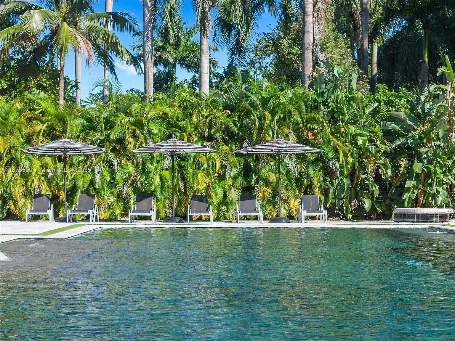 This image showcases a luxurious pool area. Lounge chairs with striped umbrellas are neatly arranged by the pool, which reflects the surrounding lush greenery. The area exudes a sense of relaxation and privacy, making it an ideal outdoor space for leisure and entertainment.