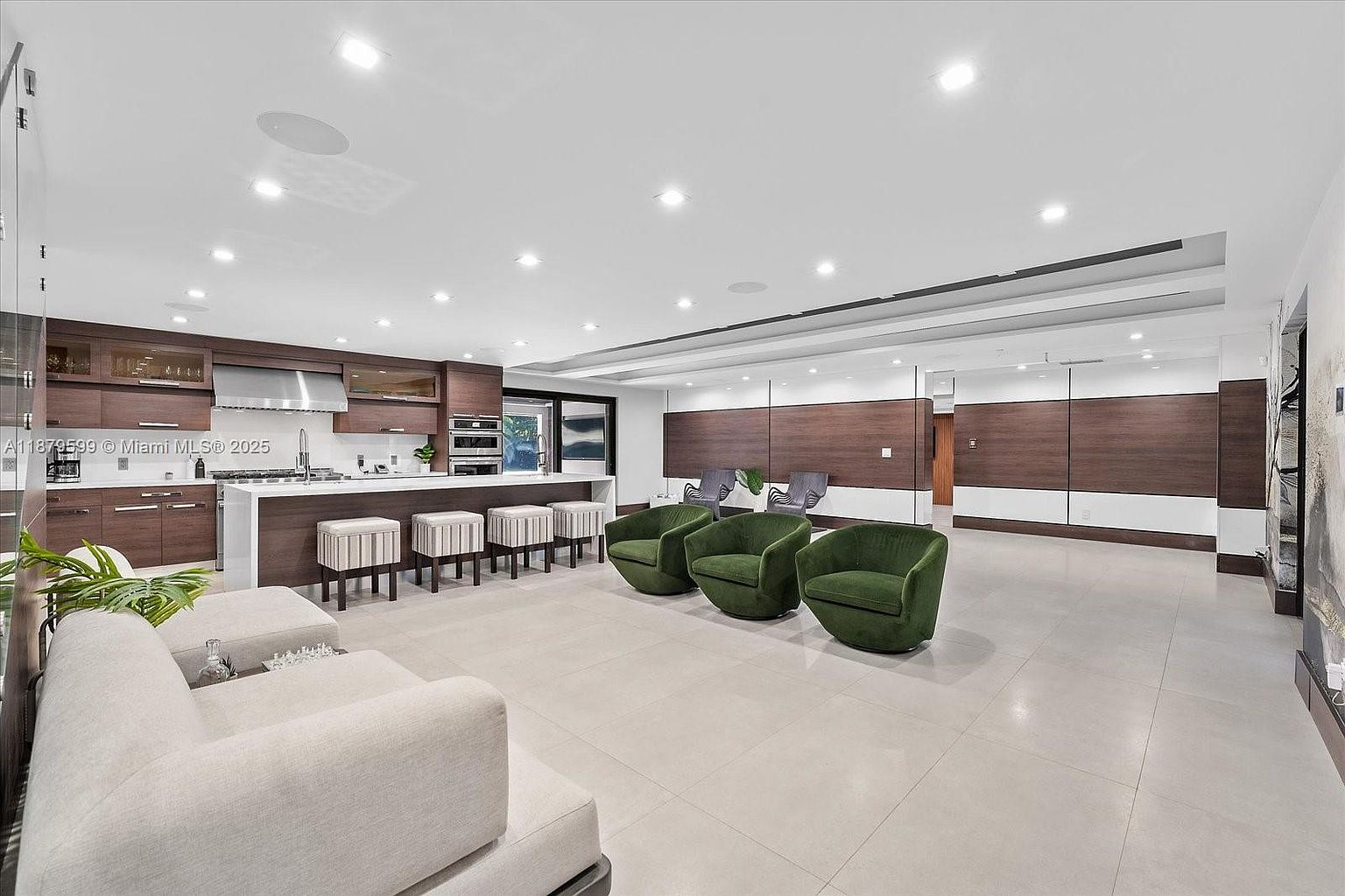 This interior shot showcases a modern living room and kitchen area. The space features a sleek kitchen with dark wood cabinetry and a white countertop island, complemented by a living area with green armchairs and a light-colored sectional sofa. The open floor plan is enhanced by recessed lighting and large format tile flooring, creating a bright and spacious atmosphere.