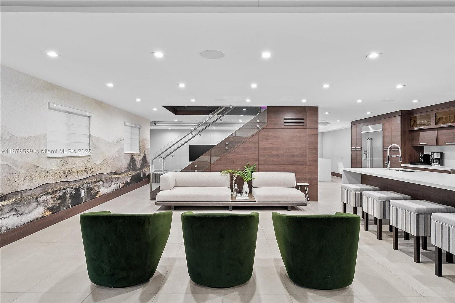 This interior shot showcases a modern living space with a seamless blend of textures and materials. The room features three dark green velvet chairs in the foreground, facing a pair of light-colored sofas separated by a small table with greenery. A striking wall mural adds depth, while the adjacent kitchen area with bar stools and sleek cabinetry suggests an open-concept design, all under recessed lighting.