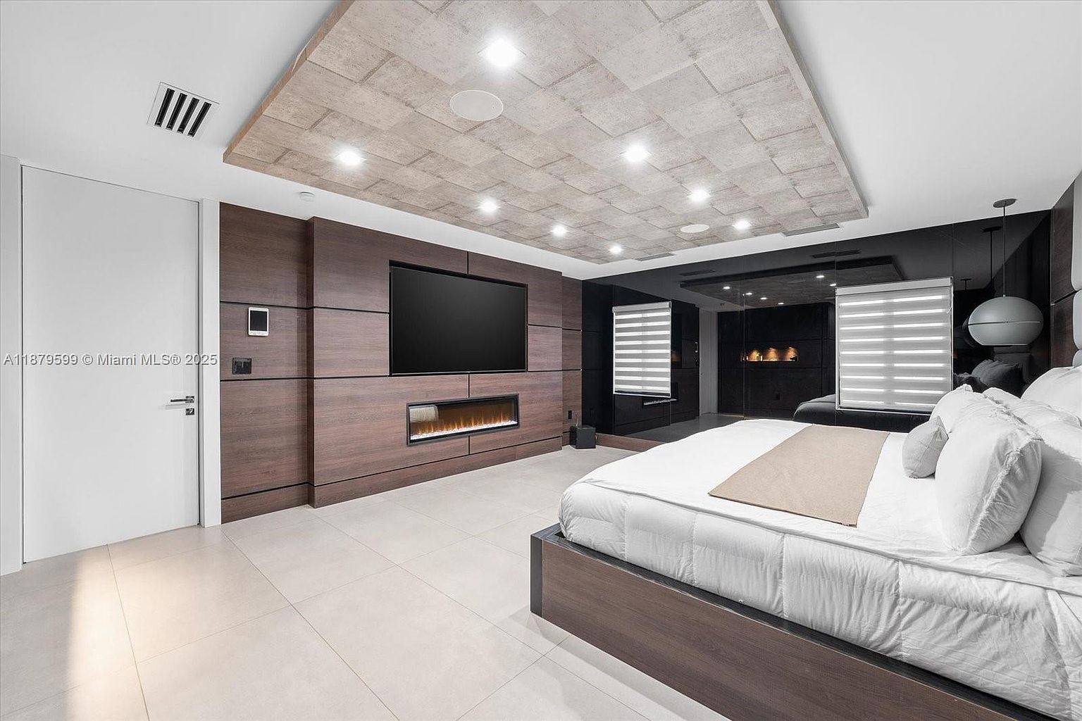 This is a primary bedroom featuring a modern design with a large bed, a wooden accent wall with a built-in fireplace and TV, and a unique textured ceiling with recessed lighting. The room has a sleek and minimalist aesthetic, with a focus on clean lines and neutral colors, creating a luxurious and comfortable atmosphere.