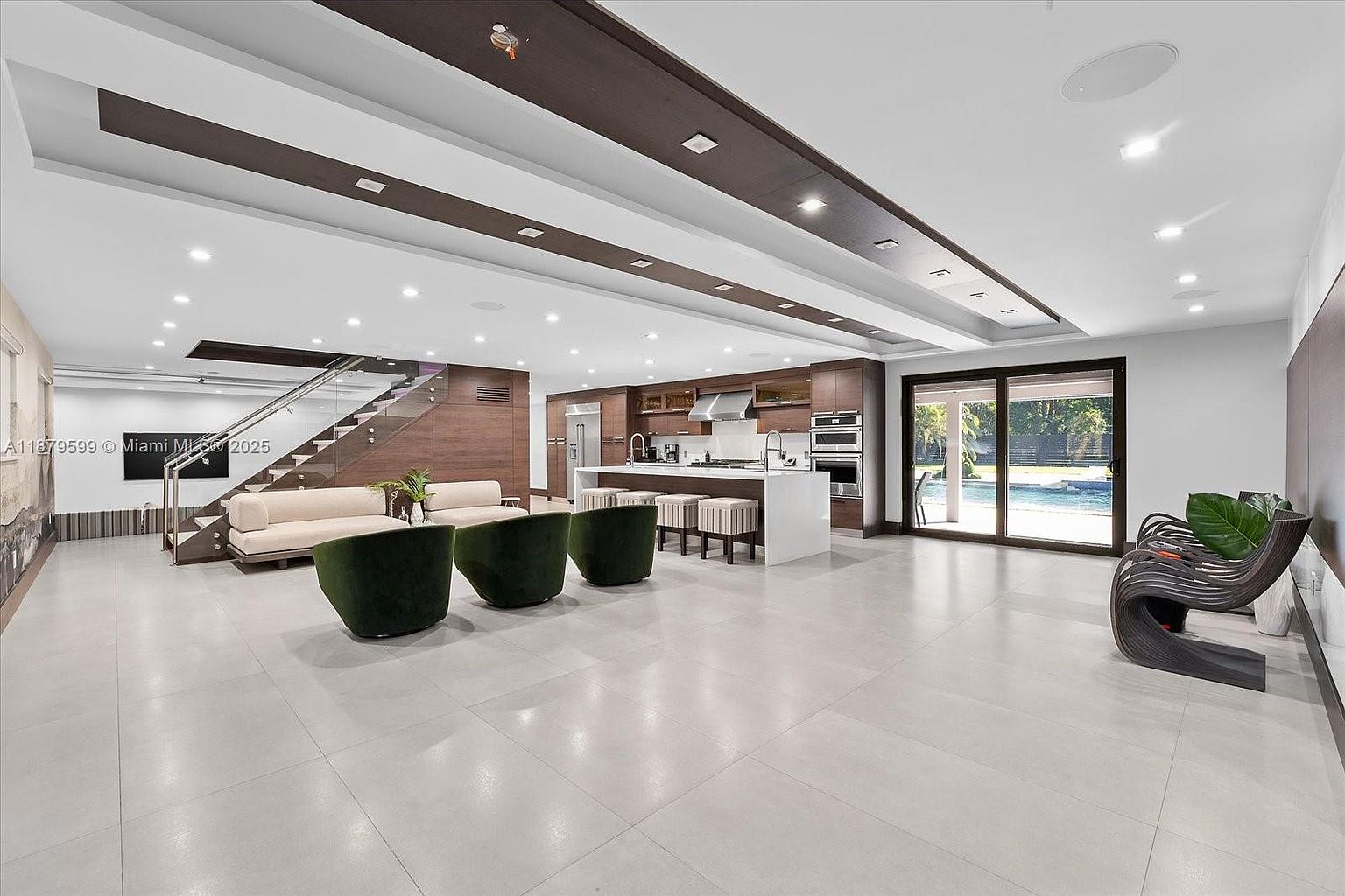 This is a spacious, modern living area that seamlessly integrates with the kitchen. The open floor plan features sleek tile flooring, contemporary furniture including green armchairs and beige sofas, and a striking staircase with glass railings. The kitchen boasts a large island and high-end appliances, while sliding glass doors provide access to the outdoor pool area, creating a luxurious and inviting atmosphere.