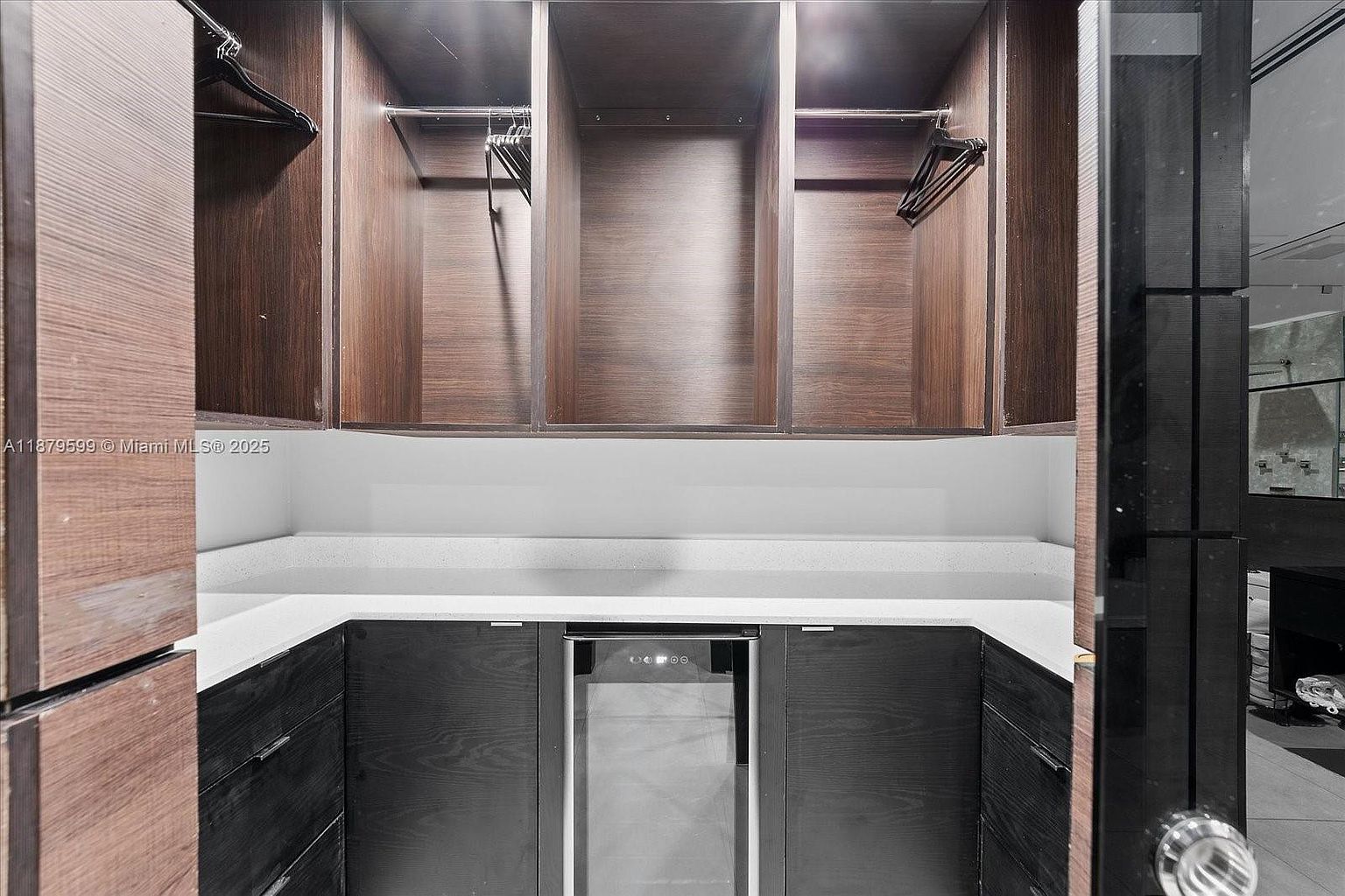 This image showcases a well-organized walk-in closet with dark wood cabinetry and a white countertop. The closet features hanging rods and open shelving, providing ample storage space. A built-in wine cooler is also visible, adding a touch of luxury.
