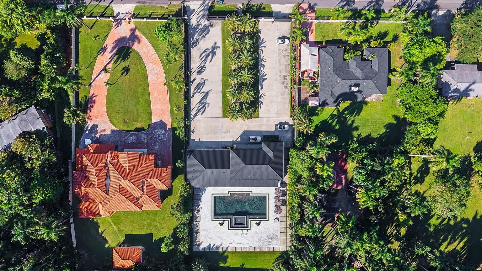 This aerial view showcases three distinct properties exhibiting luxurious features. The property on the left includes a red-tiled roof and a sprawling, circular driveway. Central to the image is a modern home with a dark roof, an immaculate square pool, and a driveway lined with palm trees, enhancing the property's curb appeal.