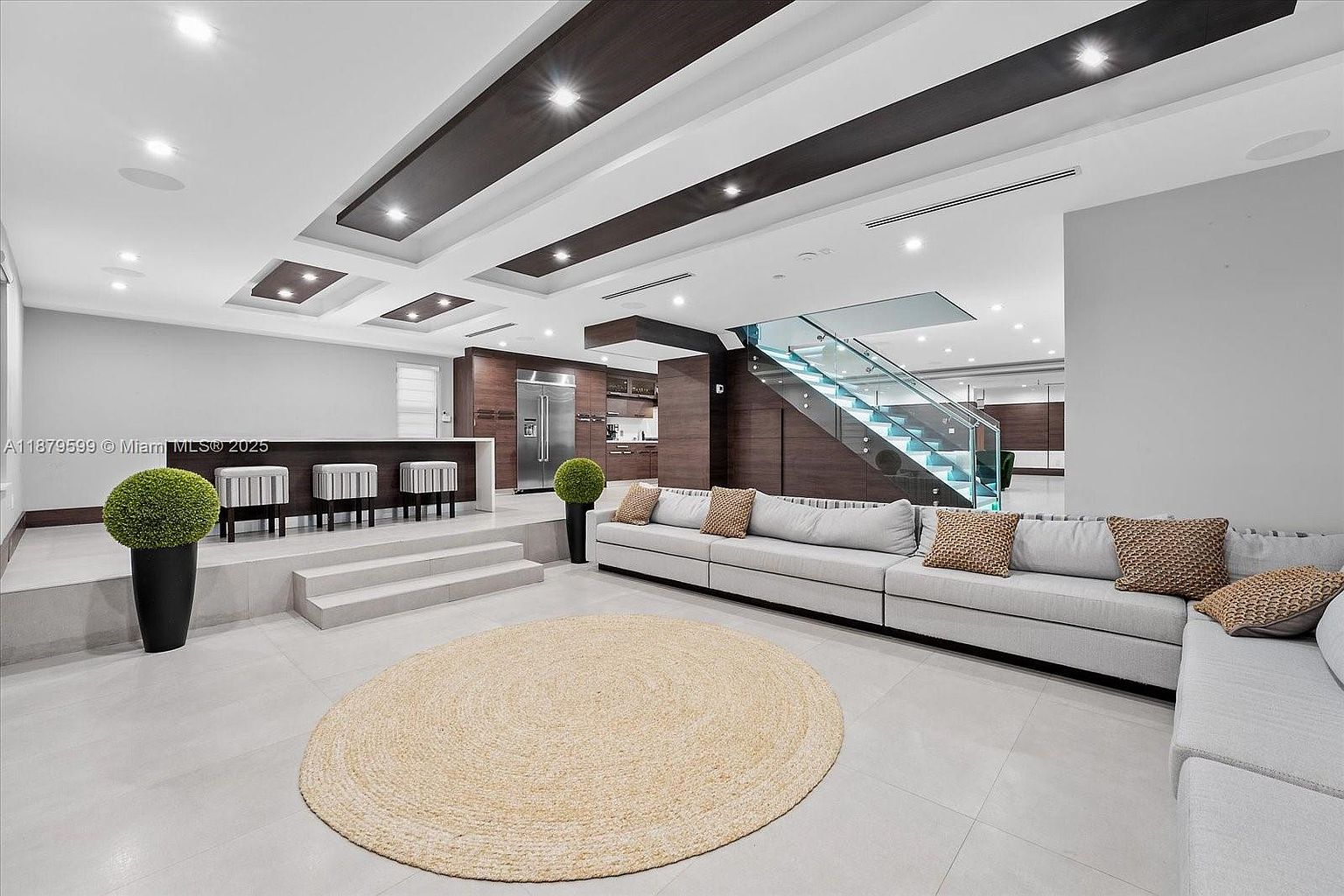 This is a spacious and modern living room featuring a large sectional sofa with decorative pillows, a round jute rug, and a bar area with stools. The room has a contemporary design with recessed lighting, dark wood accents on the ceiling, and a glass-railed staircase leading to another level. The overall impression is luxurious and inviting.