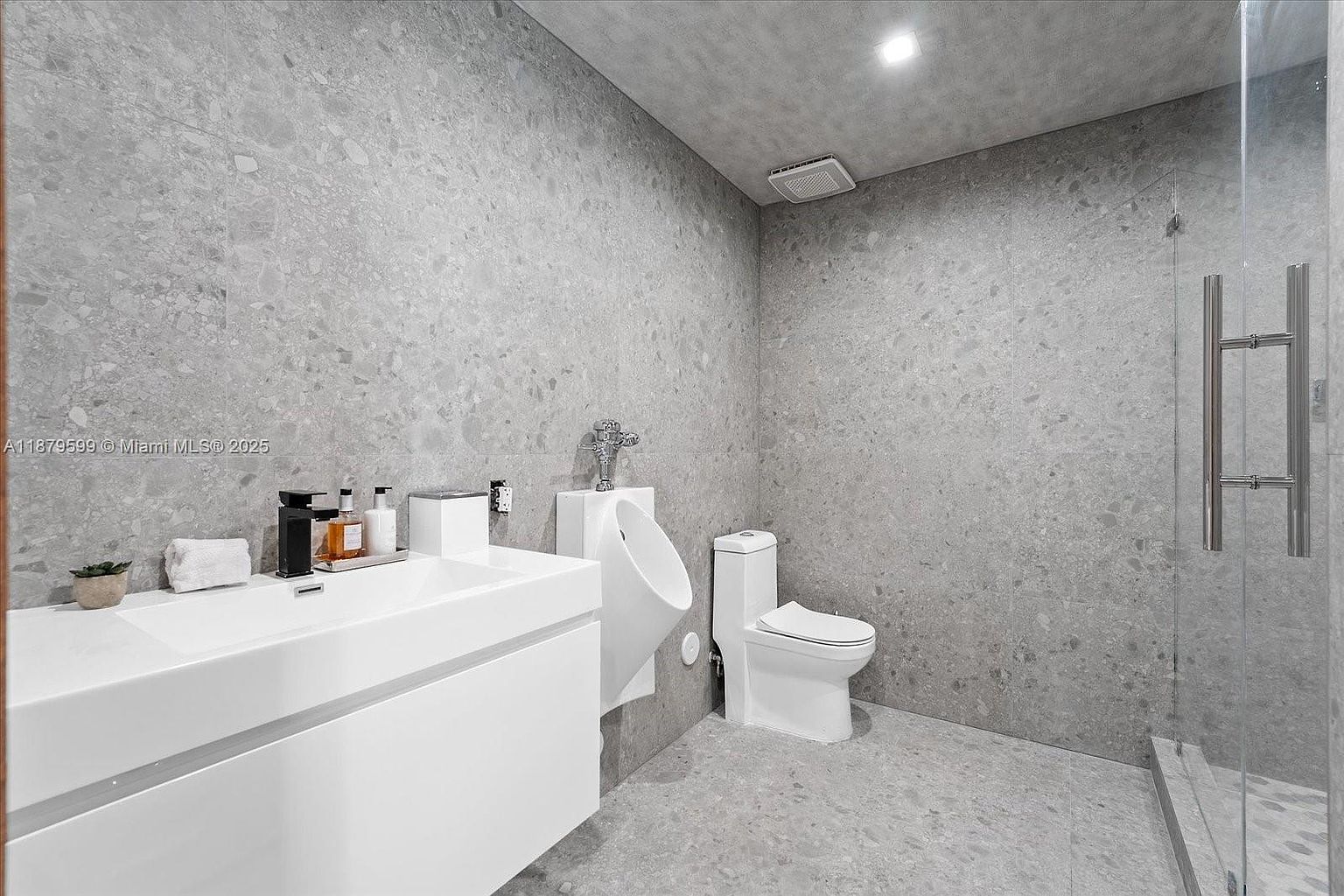 This is a modern bathroom featuring a sleek, minimalist design with gray tiled walls and flooring. The bathroom includes a white floating vanity with a rectangular sink, a wall-mounted urinal, a toilet, and a glass-enclosed shower. The overall impression is clean, contemporary, and luxurious.