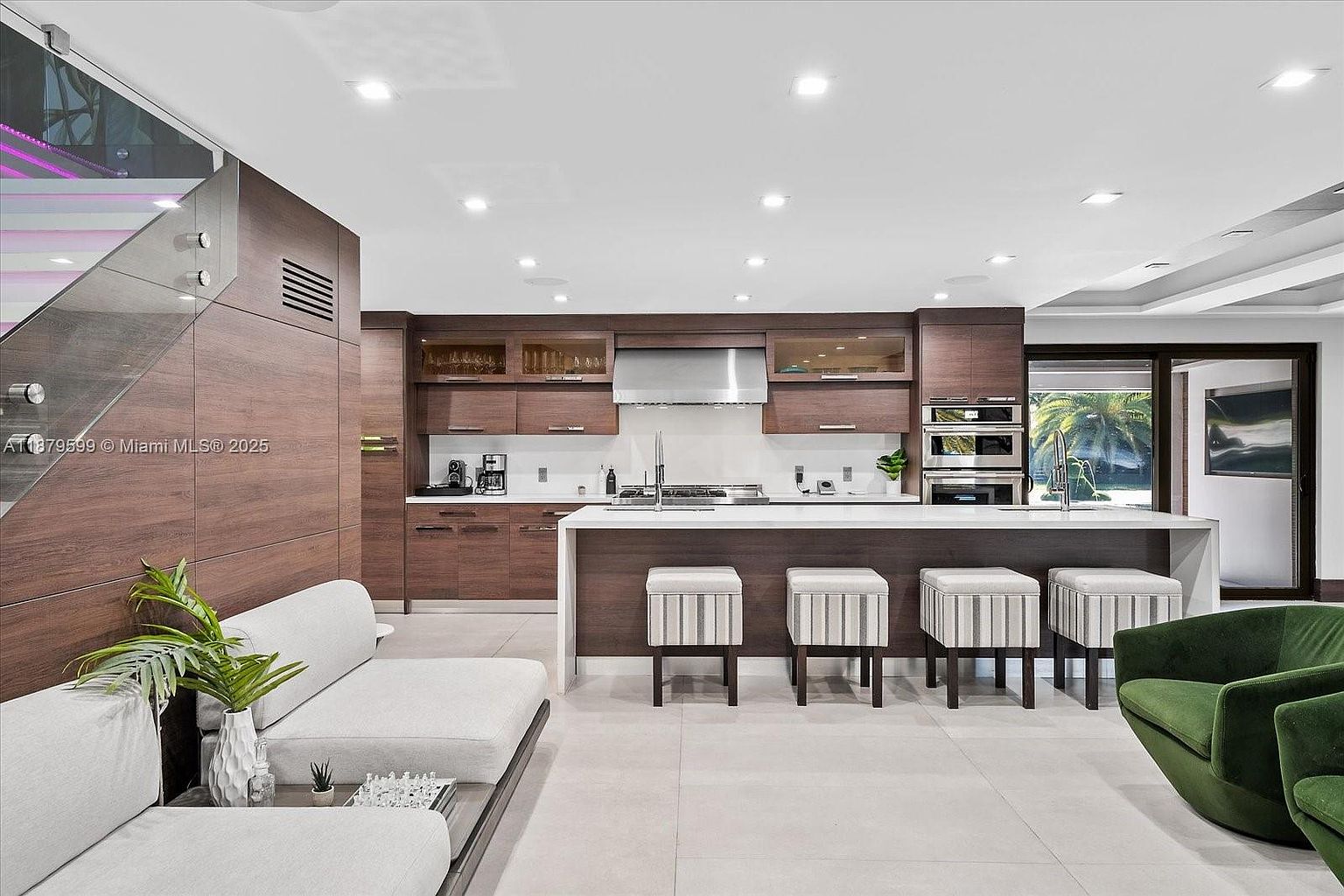 This is a modern kitchen with dark wood cabinetry, stainless steel appliances, and a large white countertop island with seating. The kitchen seamlessly blends into a living area with a light-colored sofa and green armchairs, creating an open and inviting space. A staircase with glass railings is visible on the left, adding to the contemporary design.