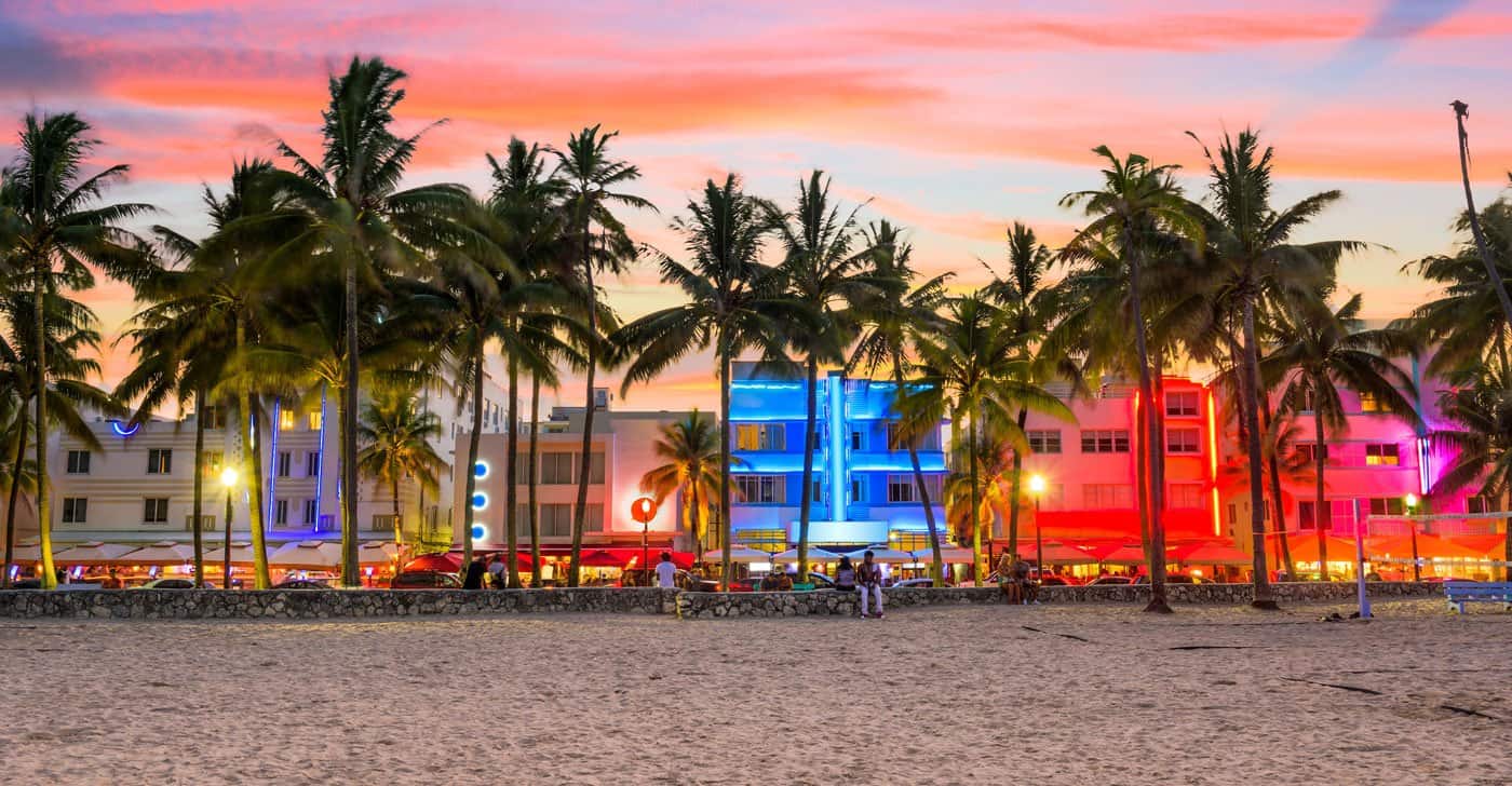 This image captures a vibrant beachfront scene featuring colorful Art Deco buildings framed by numerous palm trees under a striking pink and orange sky. The photograph highlights the lively atmosphere of Miami Beach with detail, presenting an alluring setting that emphasizes location and vibrant architecture, enhancing the appeal for potential real estate interest.