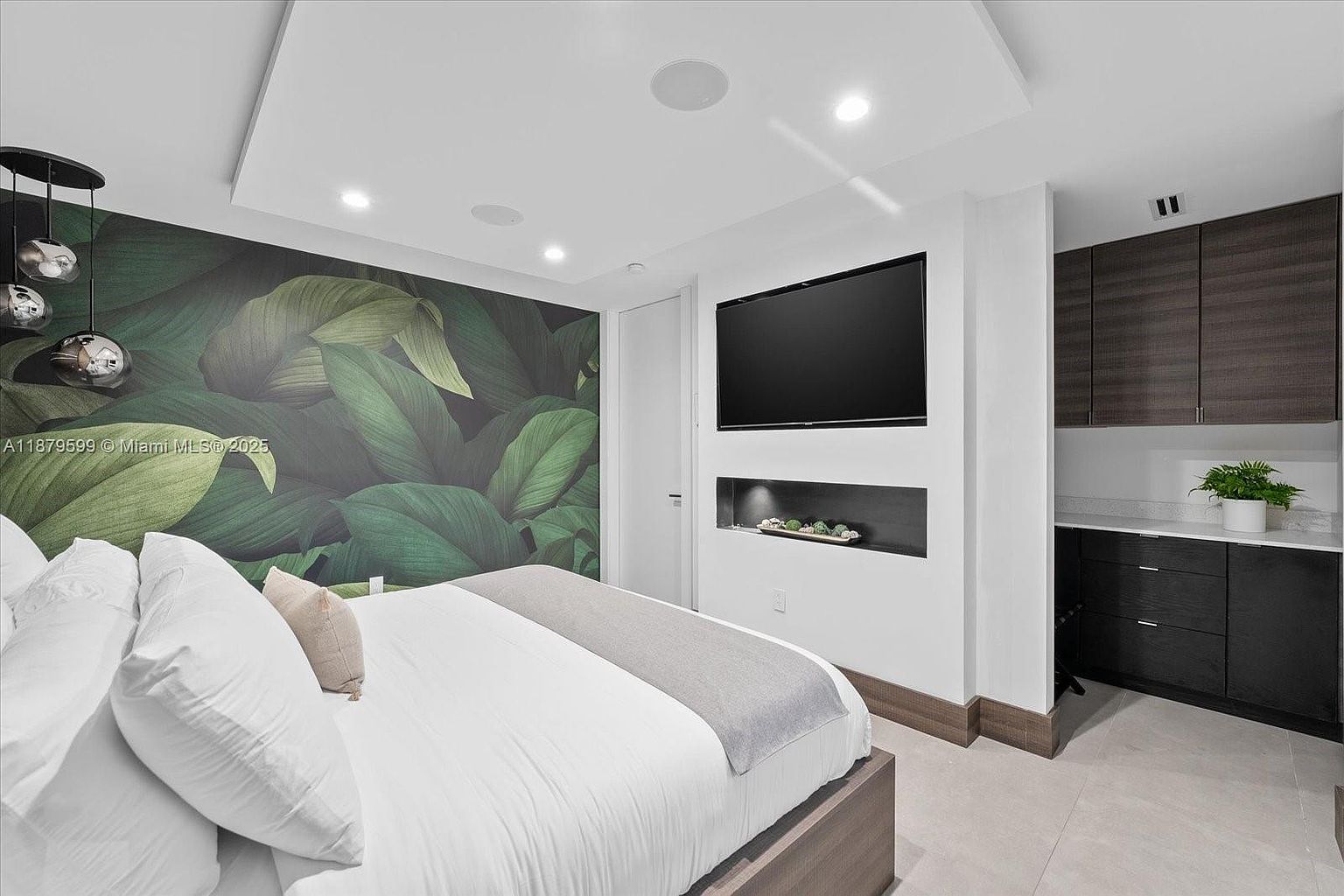 This is a primary bedroom featuring a king-size bed with white linens and a gray throw. The room has a modern design with a large flat-screen TV mounted on the wall above a decorative niche. A striking leaf-patterned wallpaper adds a touch of nature, while dark wood cabinetry provides storage and a small countertop area.