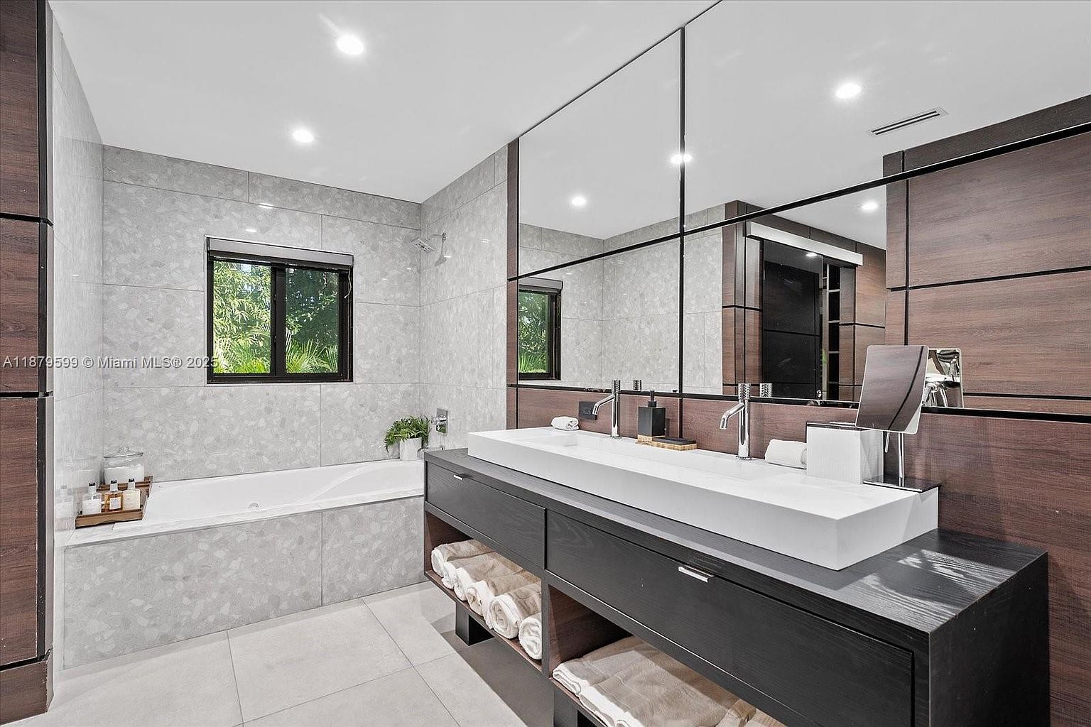 This is a primary bathroom featuring a double sink vanity with a modern, rectangular white basin and dark wood cabinetry. A large, multi-panel mirror hangs above the vanity, reflecting the natural light from a nearby window and the tiled walls. The bathroom also includes a bathtub, creating a luxurious and spa-like atmosphere.