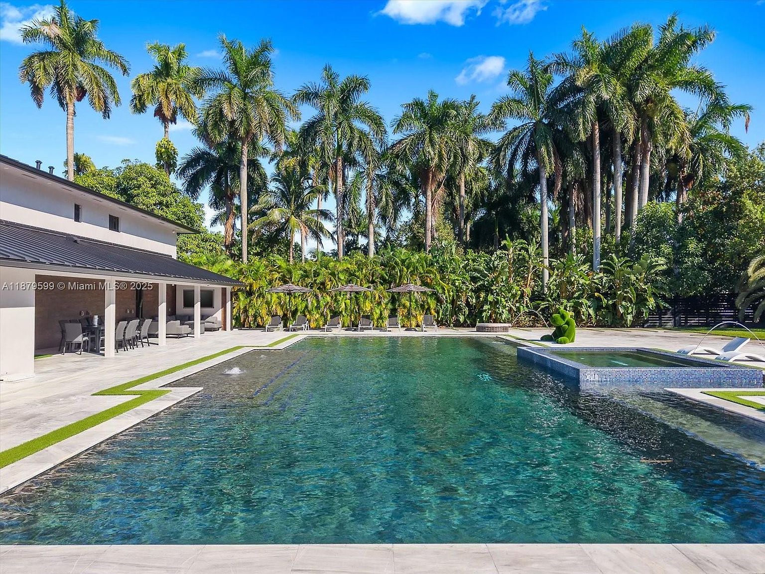 This image showcases a luxurious backyard pool and spa area. The pool features a dark, reflective surface, complemented by a smaller, tiled spa. Lush tropical landscaping and tall palm trees surround the area, creating a private and serene oasis, perfect for relaxation and entertainment.