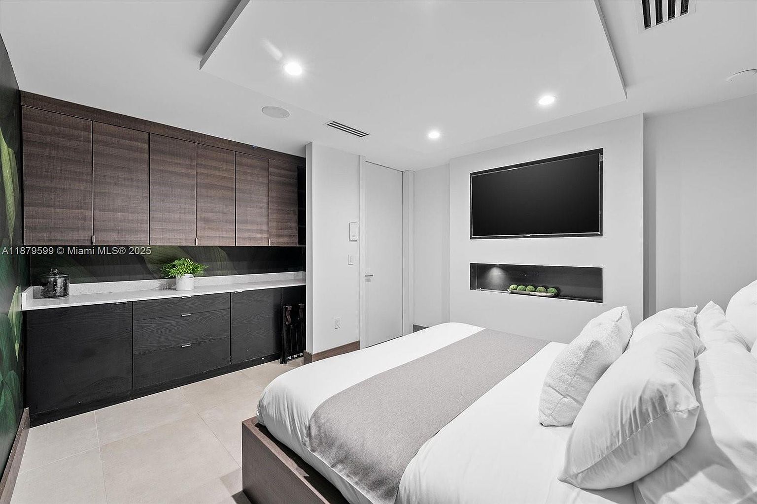 This is a primary bedroom featuring a modern design with a large bed dressed in white linens and a gray runner. A built-in entertainment center includes a large flat-screen TV and a display niche. The room also features dark wood cabinetry and a light-colored tile floor, creating a sleek and comfortable atmosphere.