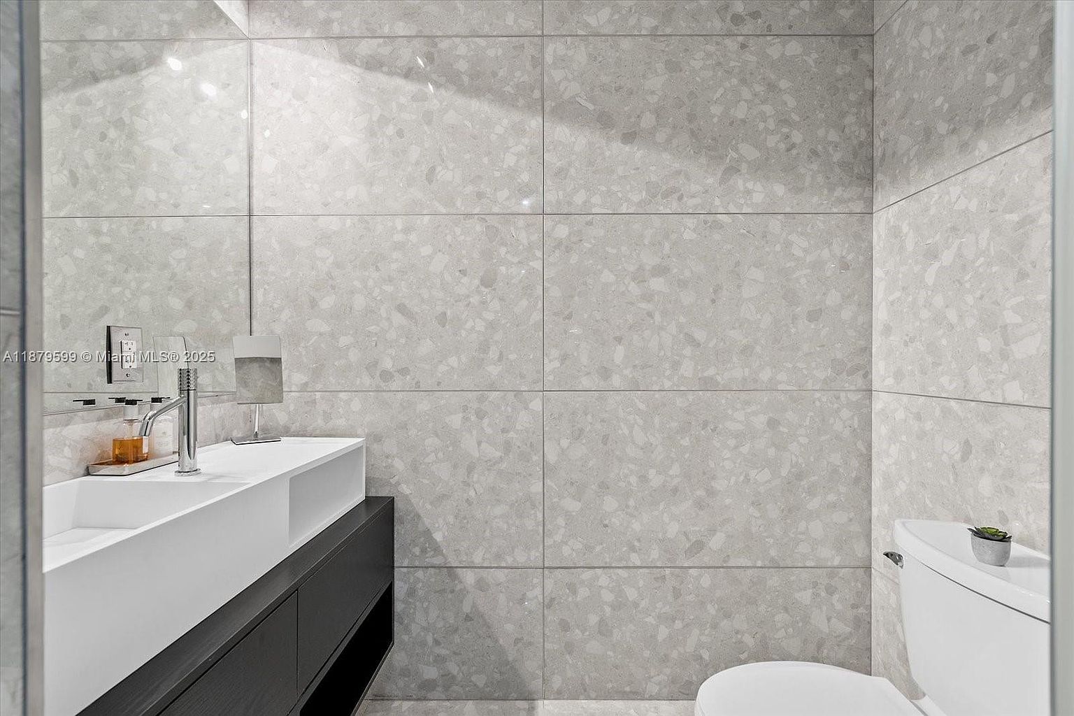 This is a modern bathroom featuring a sleek, rectangular white sink mounted on a dark wood vanity. The walls are covered in large, light gray terrazzo tiles, creating a clean and contemporary look. A toilet is visible to the right, and the overall impression is one of minimalist elegance and functionality.