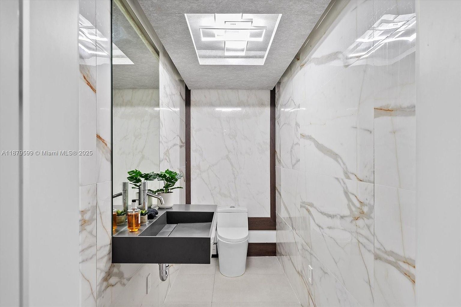 This is a modern bathroom featuring marble walls and flooring, a floating dark gray sink with a sleek faucet, and a contemporary toilet. A large mirror reflects the space, enhancing the sense of openness, while a recessed ceiling light provides ample illumination. The overall design is minimalist and luxurious.
