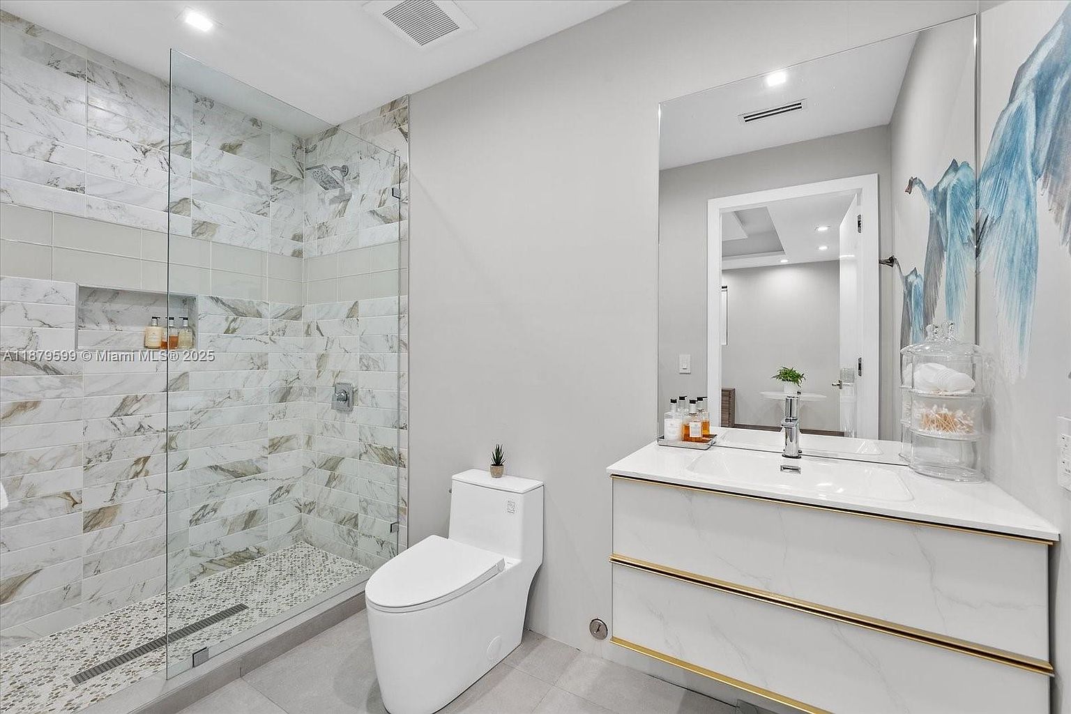 This is a modern bathroom featuring a glass-enclosed shower with marble-like tile, a white toilet, and a floating vanity with gold accents. A large mirror reflects the vanity area, and the overall design is clean and contemporary, creating a luxurious and functional space. The perspective is from the doorway, showcasing the entire bathroom.