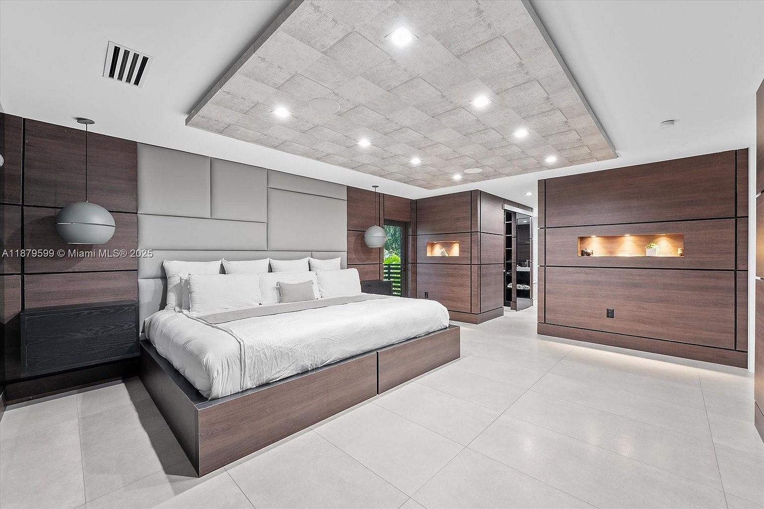 This is a luxurious primary bedroom featuring a large platform bed with white linens and a padded headboard. The room has a modern design with dark wood paneling, recessed lighting, and a unique textured ceiling. There are built-in shelves and a walk-in closet, creating a spacious and organized feel.