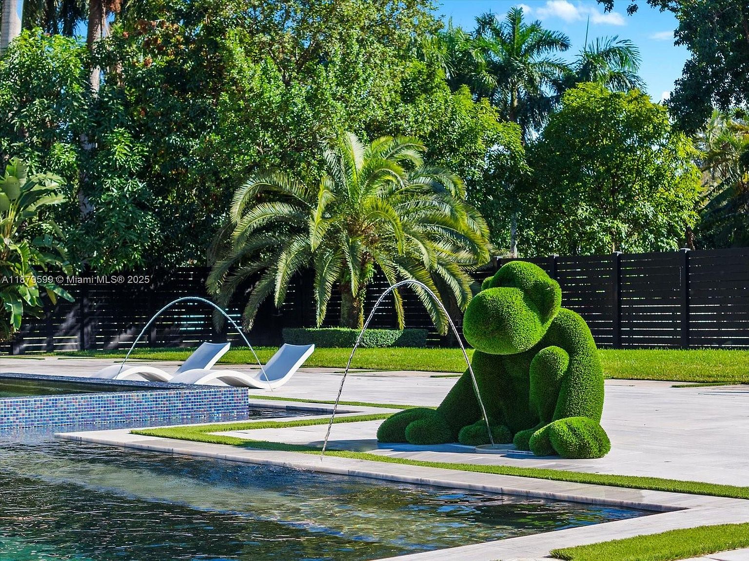 This image showcases a luxurious pool area with modern design elements. The pool features a mosaic tile edge and is surrounded by well-manicured grass and a concrete patio. A unique topiary sculpture of a gorilla sits beside the pool, adding a whimsical touch, while sleek lounge chairs and water fountains enhance the upscale ambiance.