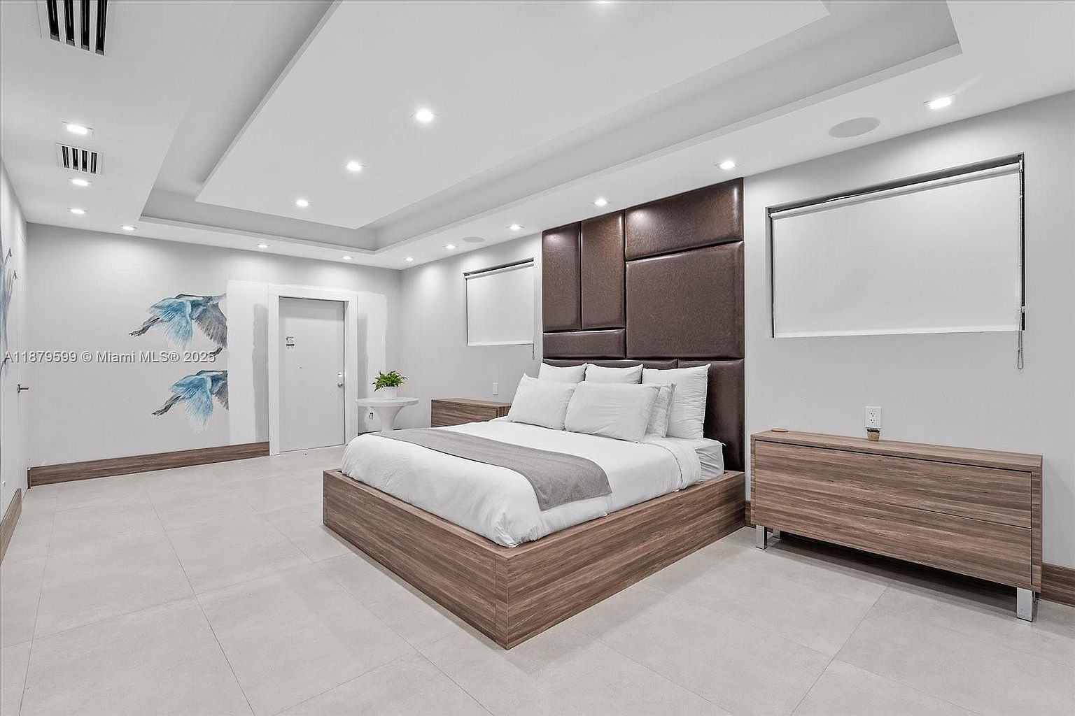 This is a primary bedroom featuring a large bed with a brown upholstered headboard. The room has recessed lighting, a modern aesthetic, and neutral color palette. There are two windows with white roller shades and matching wooden nightstands on either side of the bed.
