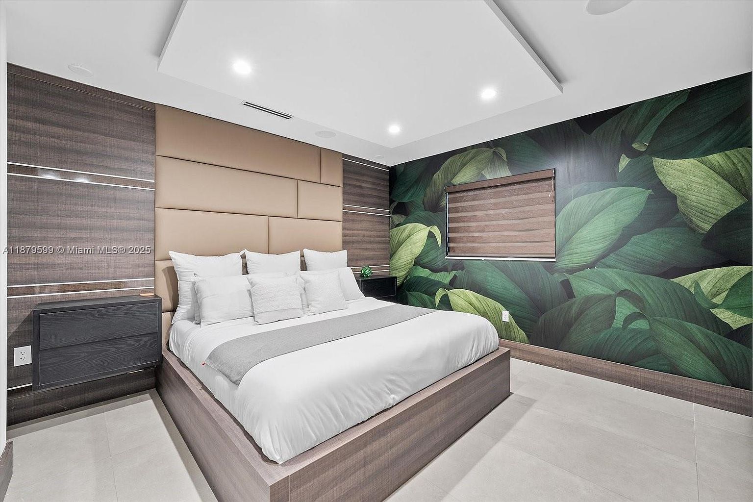 This is a primary bedroom featuring a large bed with white linens and a gray runner. The headboard is a padded, multi-panel design, flanked by wood-paneled walls. One wall is covered in a leafy green wallpaper, adding a touch of nature to the modern space. A nightstand sits beside the bed, and the room is well-lit with recessed lighting.