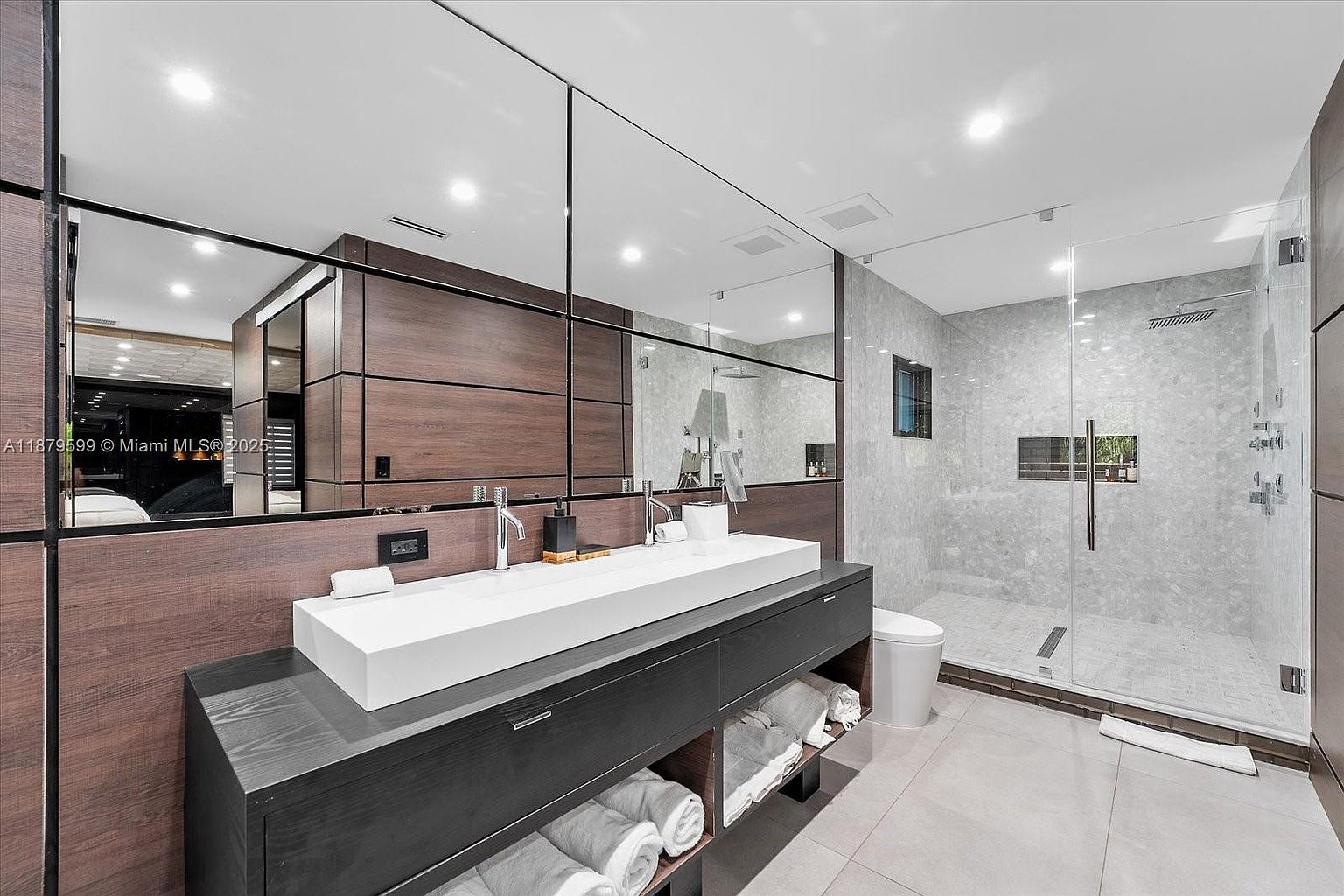 This is a modern primary bathroom featuring a long, rectangular white sink with dual faucets set on a dark gray vanity with open shelving for towels. A large mirror spans the length of the vanity, reflecting the shower area with glass doors and gray marble-like tiling. The overall impression is clean, luxurious, and well-organized.