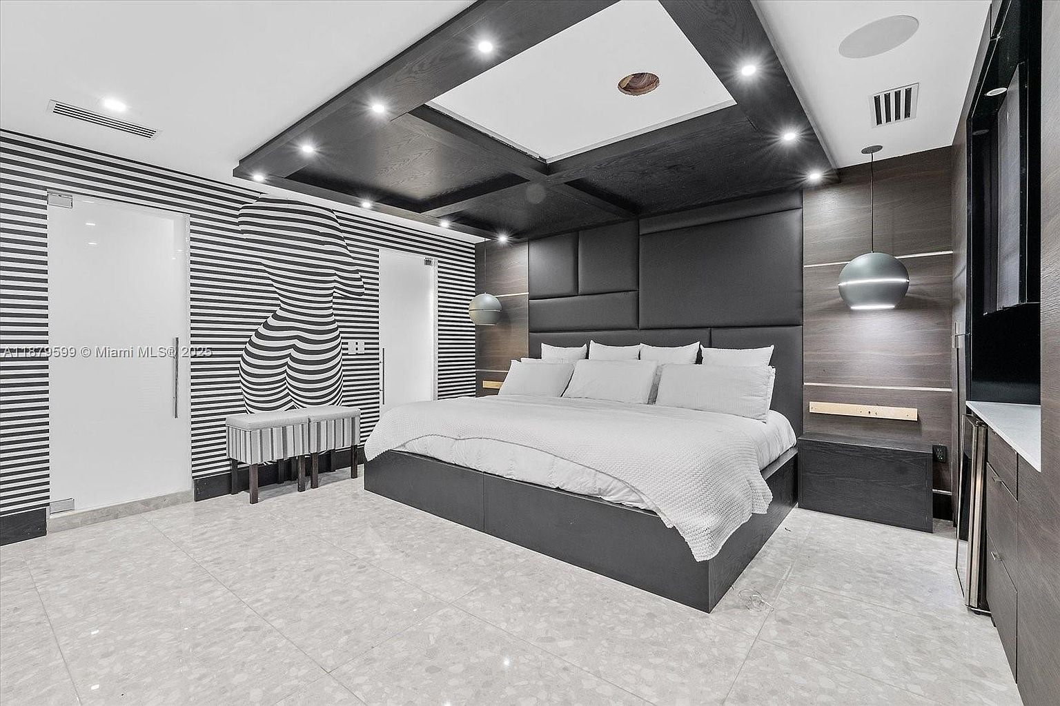 This is a modern primary bedroom featuring a large bed with a padded headboard, flanked by dark wood nightstands. The room has a unique design with striped wallpaper and a striking black wood ceiling with recessed lighting. A modern pendant light hangs over the nightstand, and the flooring is a light-colored tile.