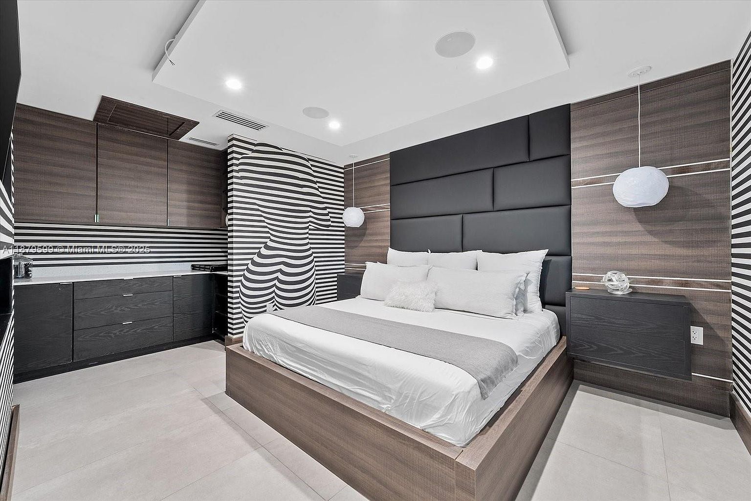 This primary bedroom features a modern design with a large bed, a padded headboard, and unique striped wall art. Built-in cabinetry and floating nightstands add to the room's sleek aesthetic. The color palette is primarily neutral, creating a calming and sophisticated atmosphere.