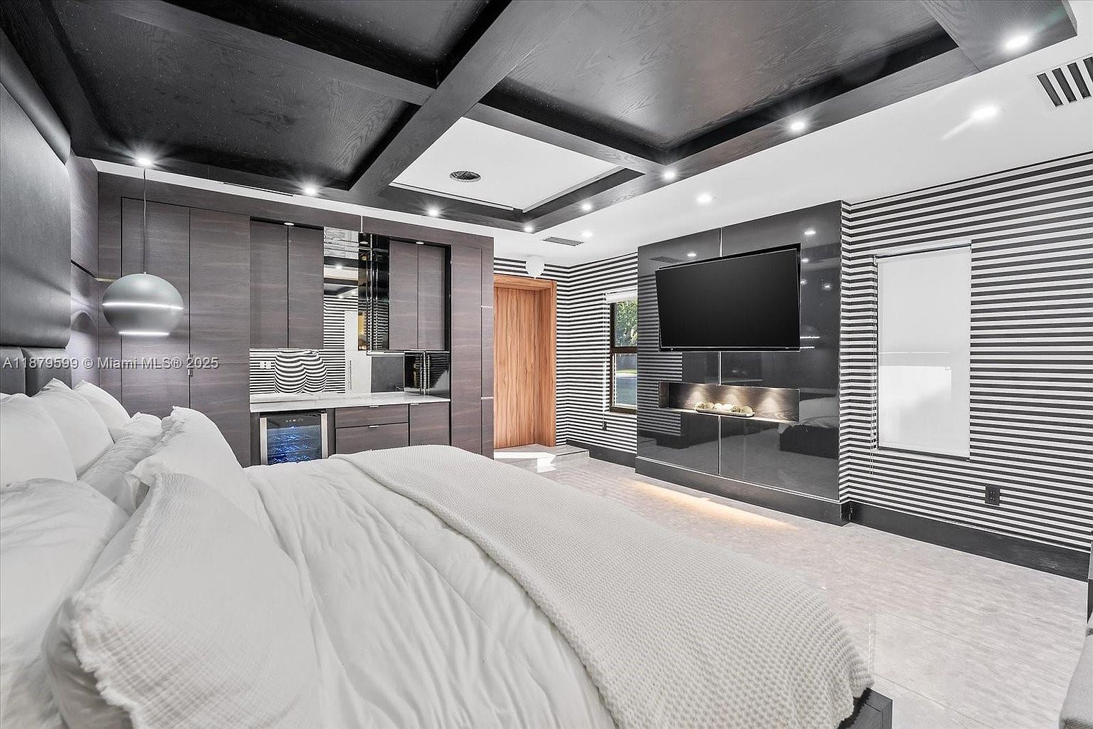 This is a primary bedroom featuring a modern design with a black and white striped accent wall and a dark wood coffered ceiling. The room includes a built-in entertainment center with a large flat-screen TV and a fireplace. A wet bar is also integrated into the design, and the bed is dressed in white linens, creating a luxurious and comfortable atmosphere.