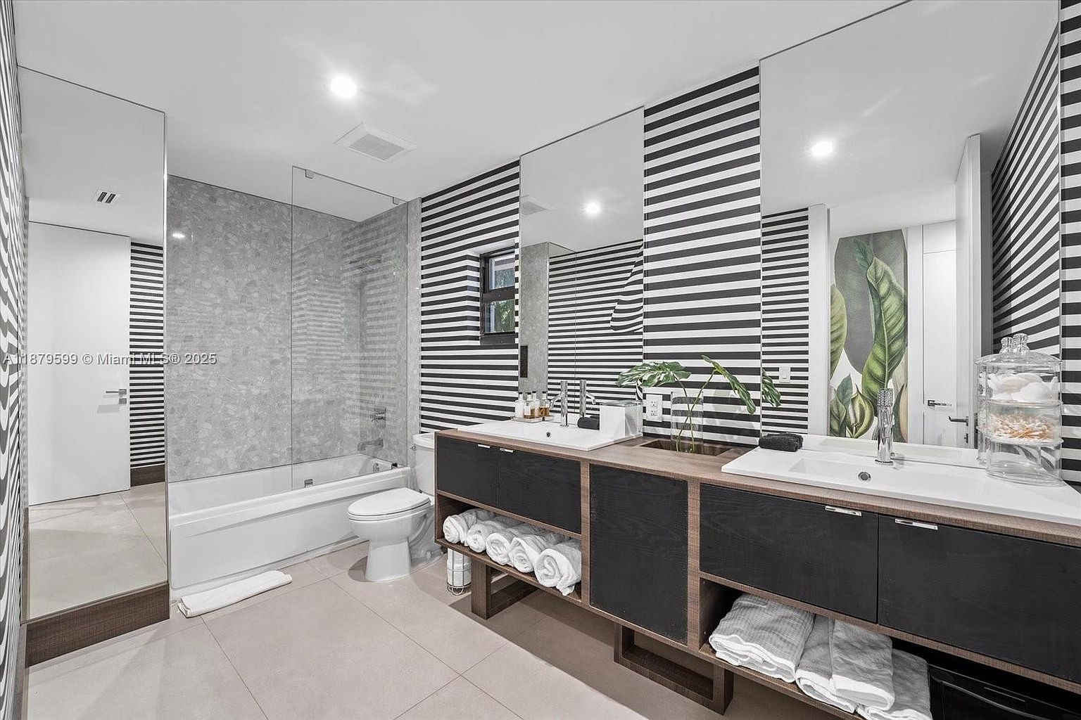 This is a modern primary bathroom featuring a double vanity with dark wood cabinetry and light countertops. The walls are adorned with a striking black and white striped pattern, complemented by large mirrors above the sinks. A glass-enclosed shower and bathtub are visible to the left, creating a luxurious and stylish space.