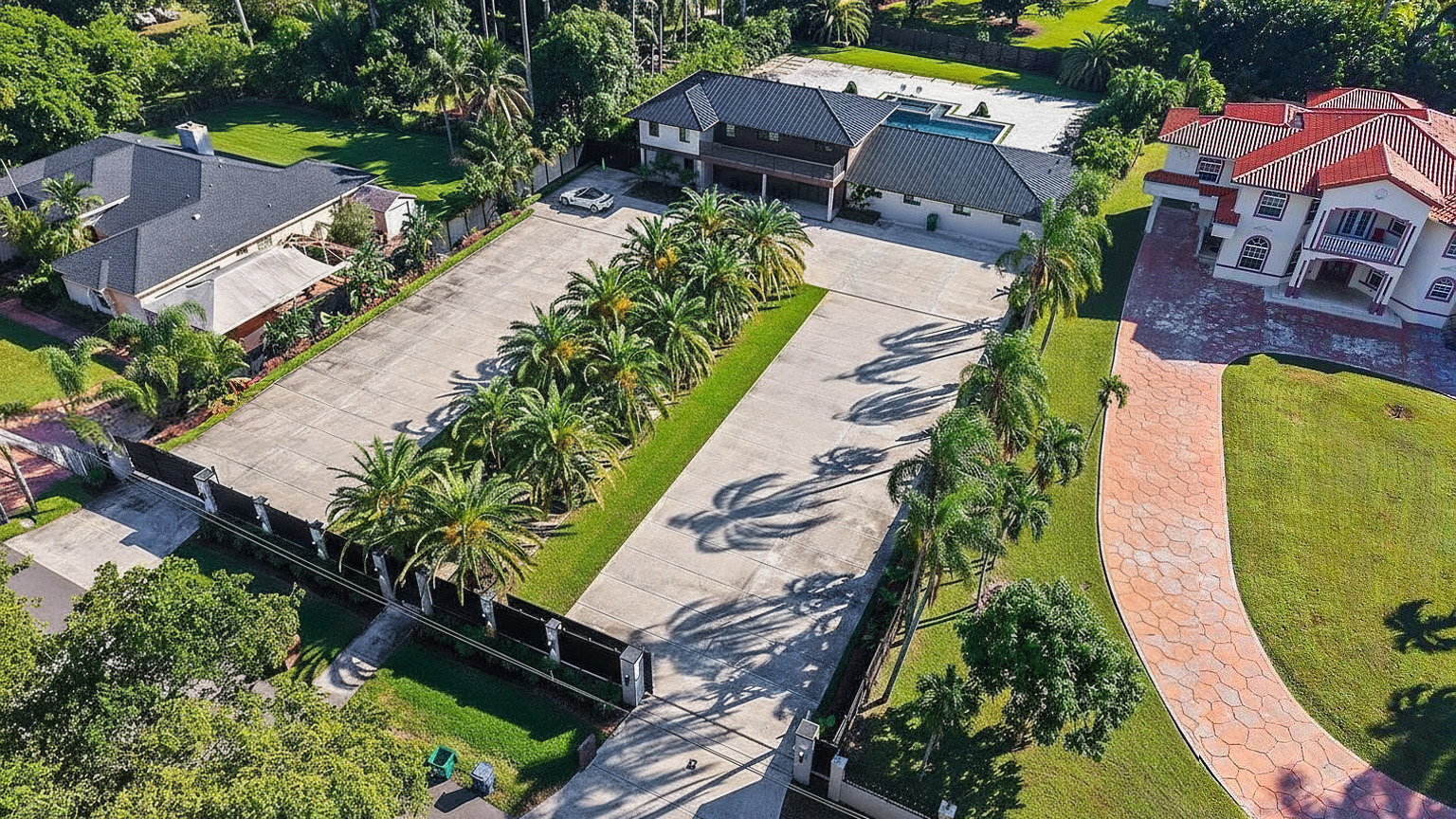 This aerial view showcases a sprawling property with multiple buildings, a large paved parking area framed by palm trees, and well-maintained landscaping. A swimming pool is visible in the background, and the overall estate presents a luxurious and private atmosphere. The driveway and walkways leading to the residence are well-defined.