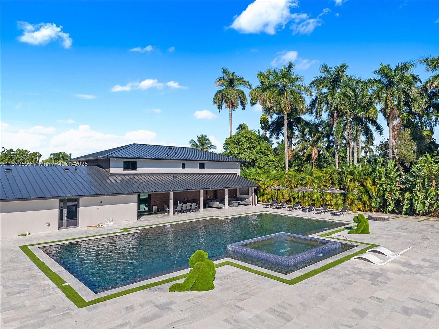 This image showcases a luxurious backyard featuring a large, dark-bottomed swimming pool with an integrated spa. The pool area is surrounded by a spacious patio made of light-colored stone tiles, complemented by manicured green turf accents. Lush tropical landscaping and tall palm trees provide privacy and enhance the resort-like atmosphere, creating an inviting outdoor living space.