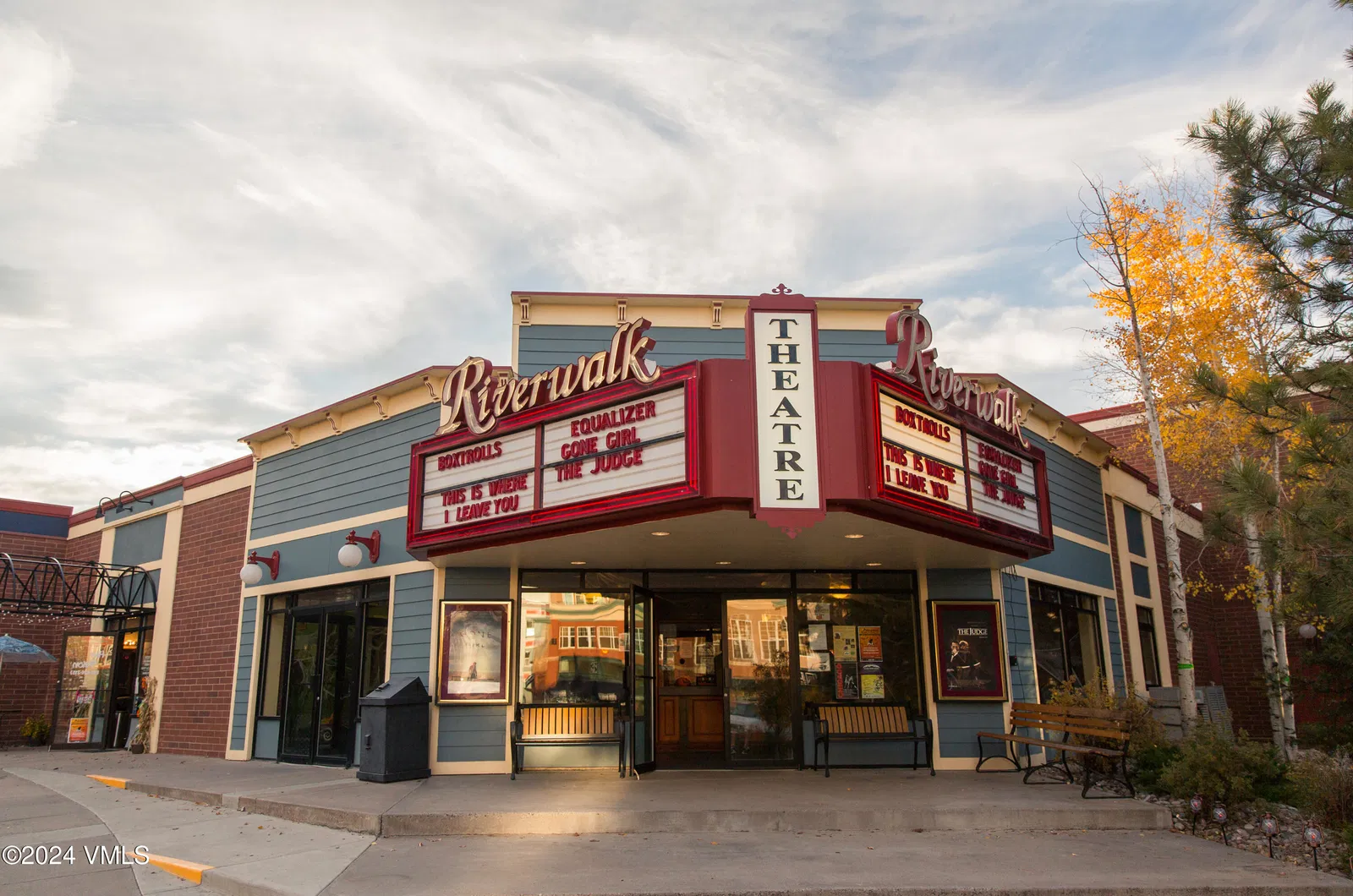 This is a front view of a Riverwalk Theater. The building has a classic facade with blue siding, brick accents, and a prominent marquee displaying movie titles. It evokes a sense of nostalgia and local charm.