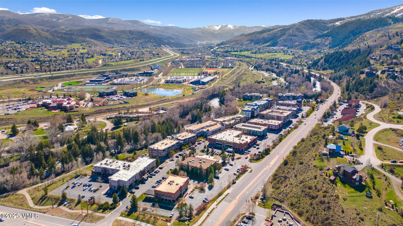 This aerial view showcases a sprawling commercial area nestled amidst a mountainous landscape with some snow still visible on the peaks. Multi-story buildings are present with ample parking. The area has a mix of commercial properties, residential areas, and green spaces.