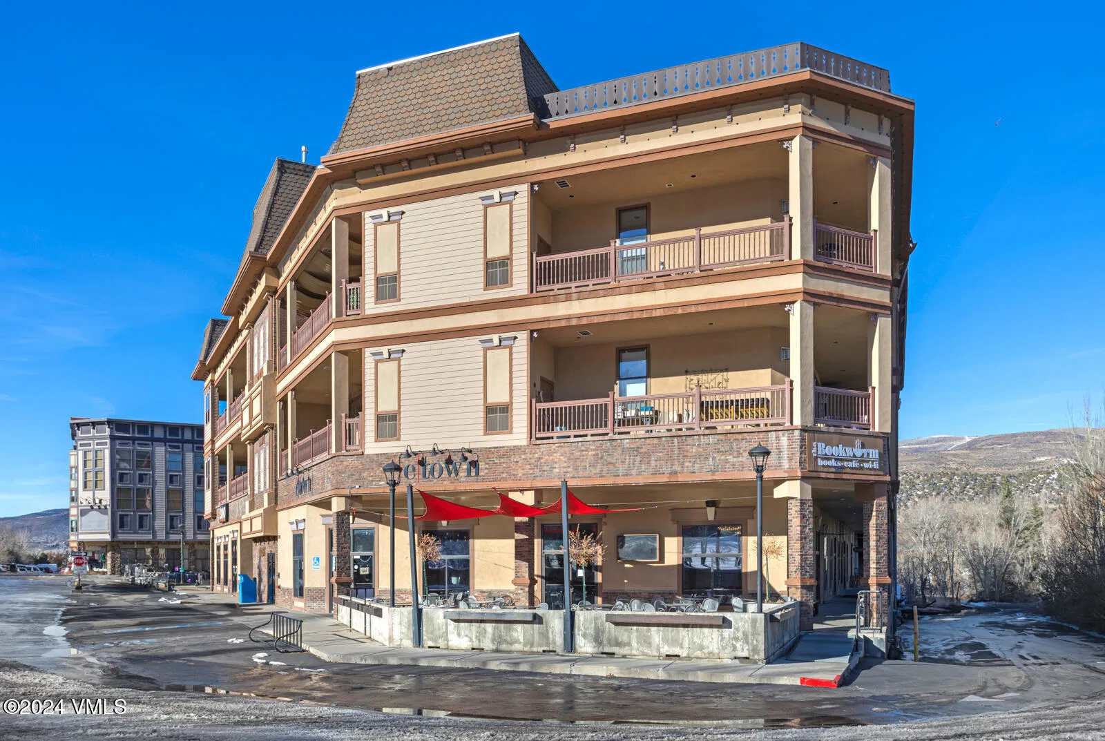 This is a front exterior view of a multi-story commercial building with residential units above. The building features a combination of tan siding and brick accents, along with well-maintained balconies. On the ground floor, a restaurant named "etoww" with red awnings offers outdoor seating, adding to the building's appeal.