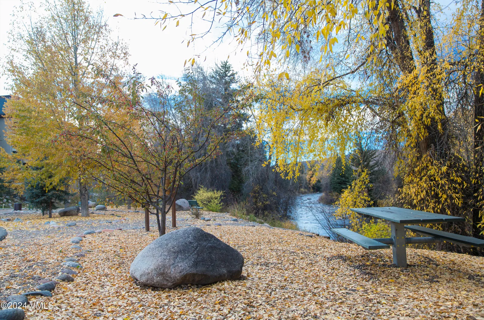 The image showcases a peaceful outdoor space in autumn, potentially part of a real estate property. A picnic table sits amid fallen leaves, offering a cozy spot for outdoor relaxation. The presence of mature trees and a nearby flowing river adds to the tranquility and natural beauty of the yard.