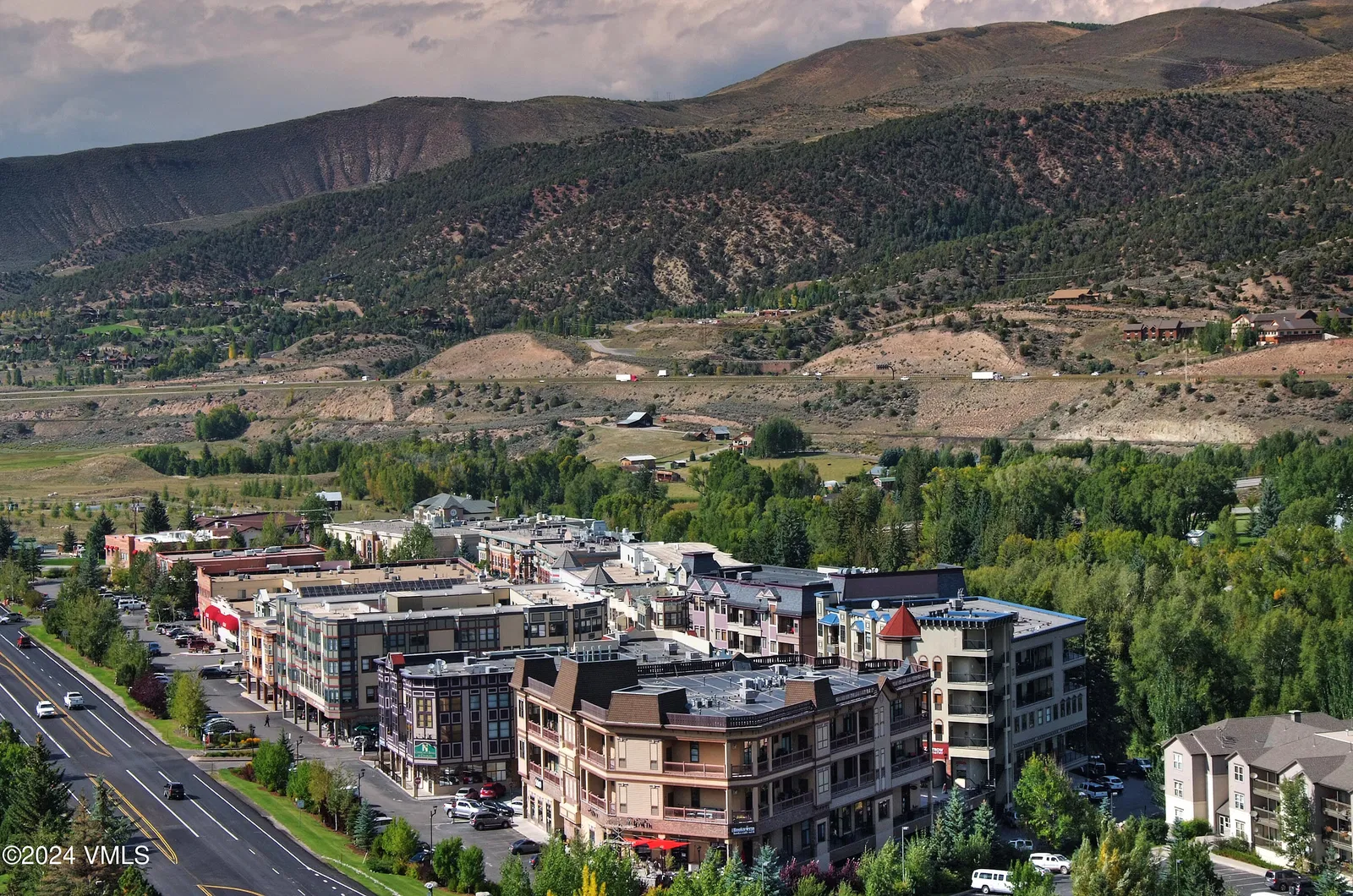 This aerial shot showcases a vibrant mountain town with multi-story buildings and a scenic backdrop of rolling hills. Lush greenery surrounds the buildings, creating a harmonious blend of urban development and natural beauty. A highway runs parallel to the town, providing easy access.