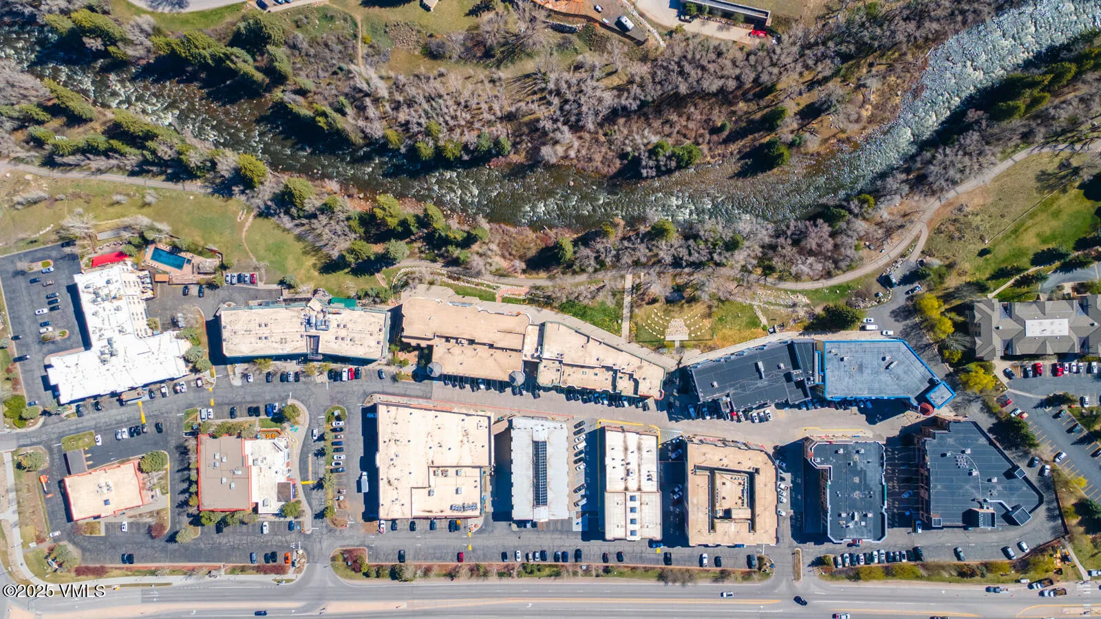 This is an aerial view of a commercial and residential area situated alongside a river. The image showcases various buildings with parking lots, trees, and a glimpse of the river winding through the landscape. The setting appears well-maintained and highlights the proximity of buildings to the natural environment.