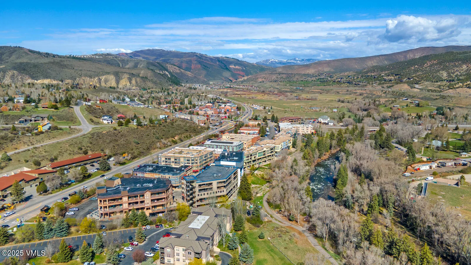 This aerial shot showcases a picturesque town nestled in a valley with mountain views. The buildings have a mix of architectural styles, featuring both residential and commercial properties. A river runs alongside the town adding natural charm and potential recreational opportunities.
