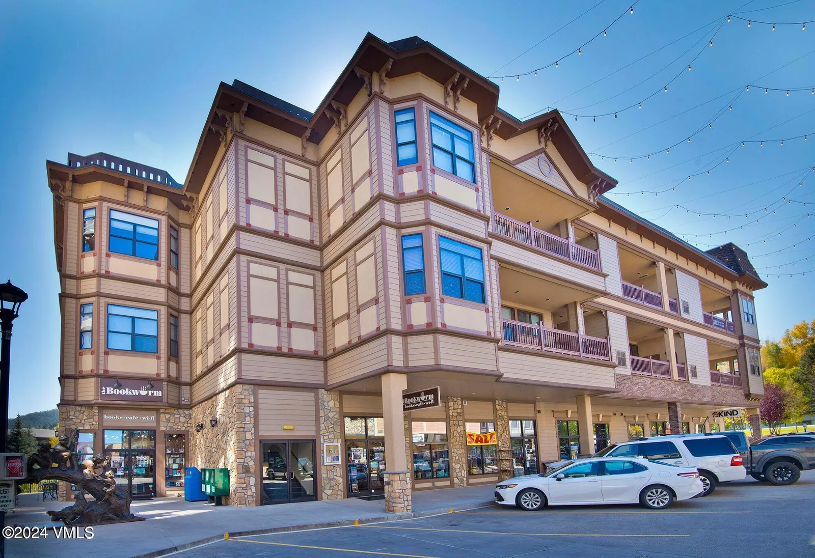 The image showcases the front exterior of a multi-story commercial building with a distinctive architectural style, featuring a combination of stone and painted siding. Prominent signage indicates retail spaces on the ground floor, suggesting a mixed-use property. Several parked cars are visible in front of the building, highlighting its accessibility and street presence.