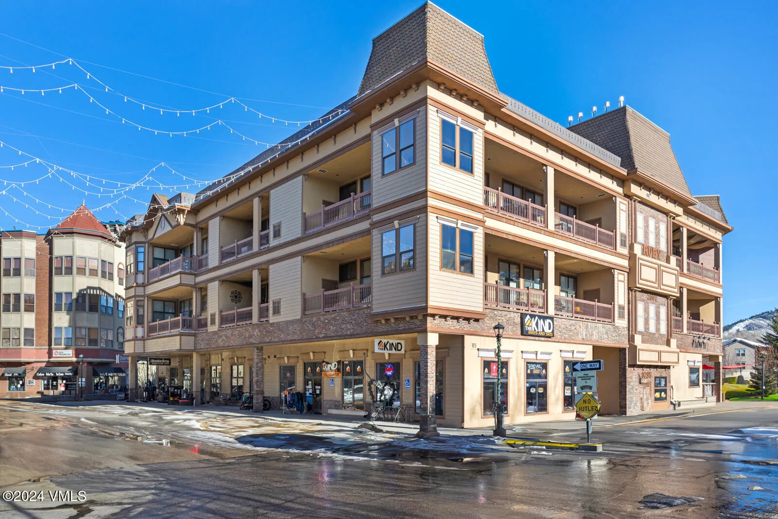 This is a front exterior view of a multi-story commercial building. The building features a combination of light brown siding and brick accents. It includes several balconies and retail spaces at the ground level, creating a mixed-use property suitable for both residential and commercial purposes.