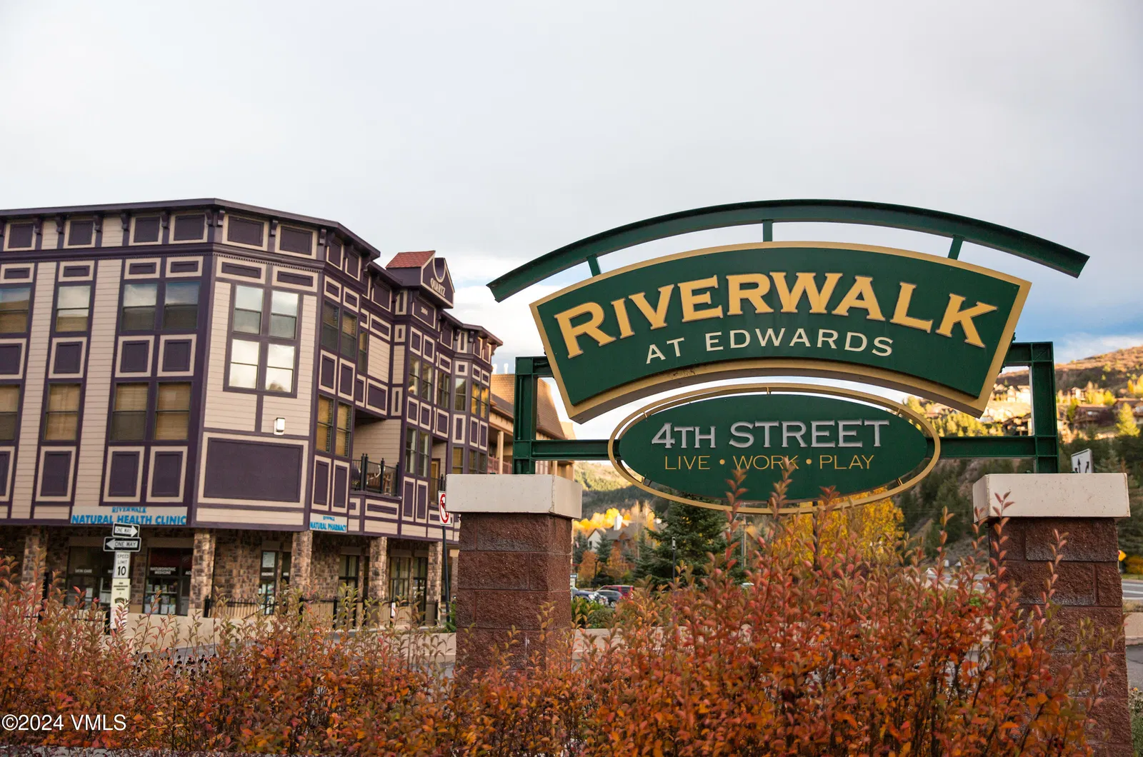 This image displays the Riverwalk at Edwards, with a prominent sign indicating the development's name and "4th Street: Live, Work, Play." The building features a unique architectural design with a combination of beige and purple accents. Mature landscaping in the foreground enhances the property's curb appeal and creates a welcoming atmosphere.