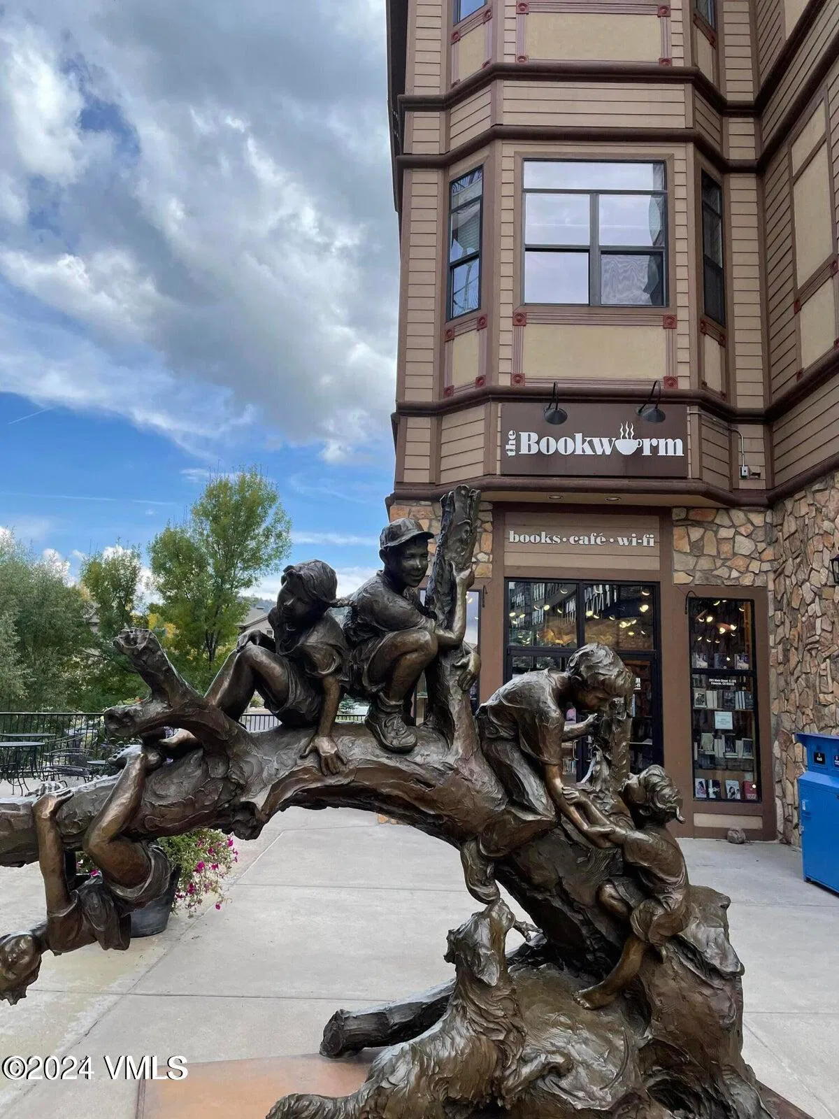 This image showcases the front view of a building with commercial space denoted by the "The Bookworm" sign and a sculpture of children climbing a tree in front of it. The building's exterior features a mix of stone and framed window designs. The presence of the sculpture adds a unique and artistic touch to the property's curb appeal.