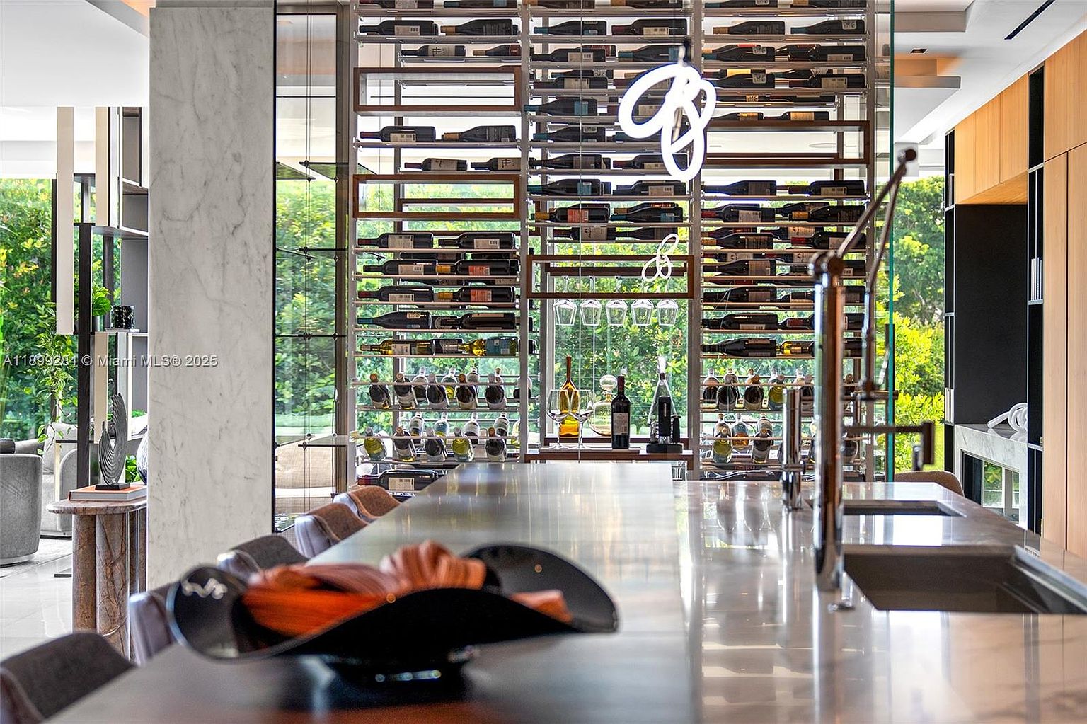 This interior shot showcases a modern kitchen with a prominent wine rack feature, filled with numerous bottles and illuminated by a decorative light fixture. A sleek countertop extends into the foreground, displaying decorative items, while the background reveals lush greenery through the windows, creating a sophisticated and inviting atmosphere.