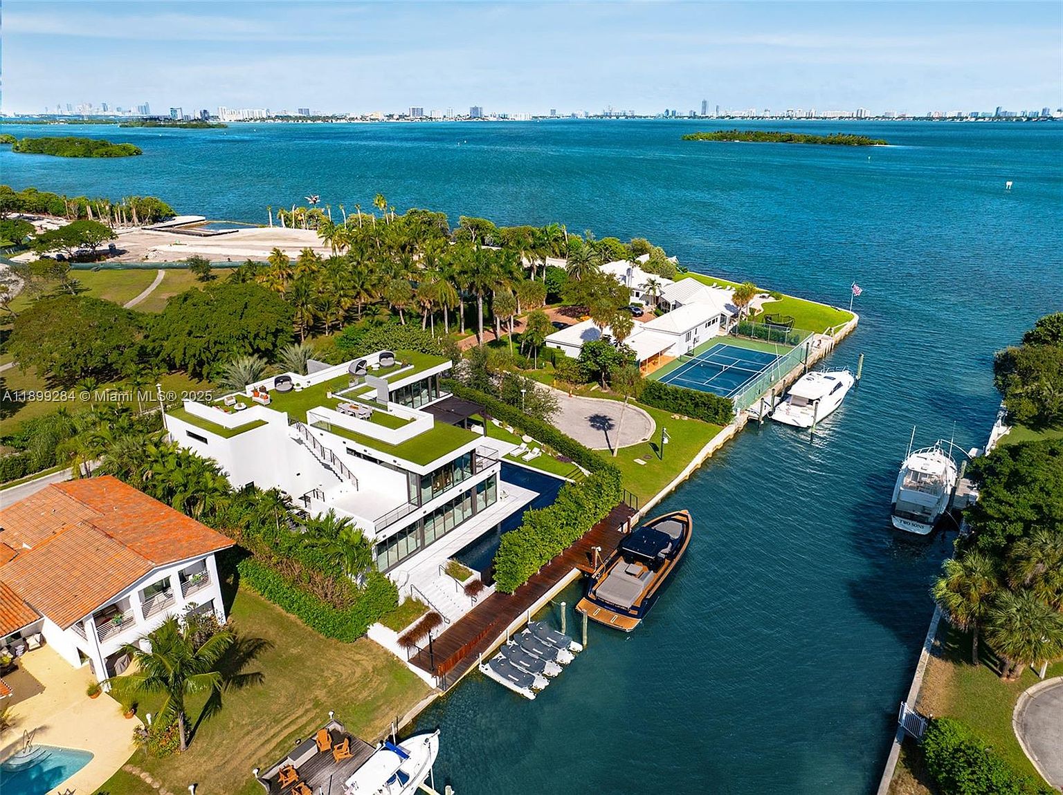 This aerial shot showcases a luxurious waterfront property featuring modern architecture with a green roof, a private dock with multiple boats, and a tennis court. The property is surrounded by lush landscaping and offers stunning views of the bay and distant city skyline. The overall impression is one of opulence and exclusivity.