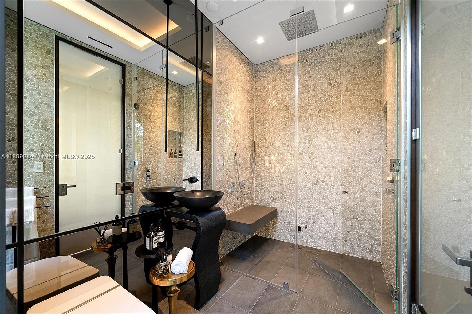 This is a modern primary bathroom featuring a double vanity with vessel sinks, a large mirror, and a walk-in shower with pebble-style tiling. The shower includes a built-in bench and a rainfall showerhead. The overall design is sleek and luxurious, with a focus on clean lines and high-end finishes.