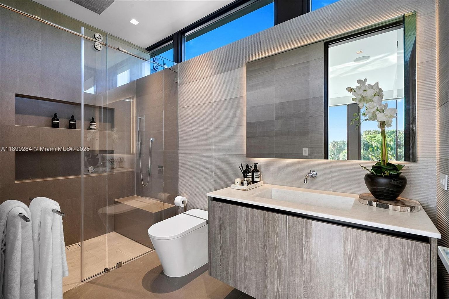 This is a modern primary bathroom featuring a glass-enclosed shower with a built-in niche, a floating vanity with a stone countertop and integrated sink, and a sleek toilet. The walls are covered in textured gray tiles, and a large mirror with integrated lighting adds to the contemporary aesthetic. A potted orchid provides a touch of elegance.