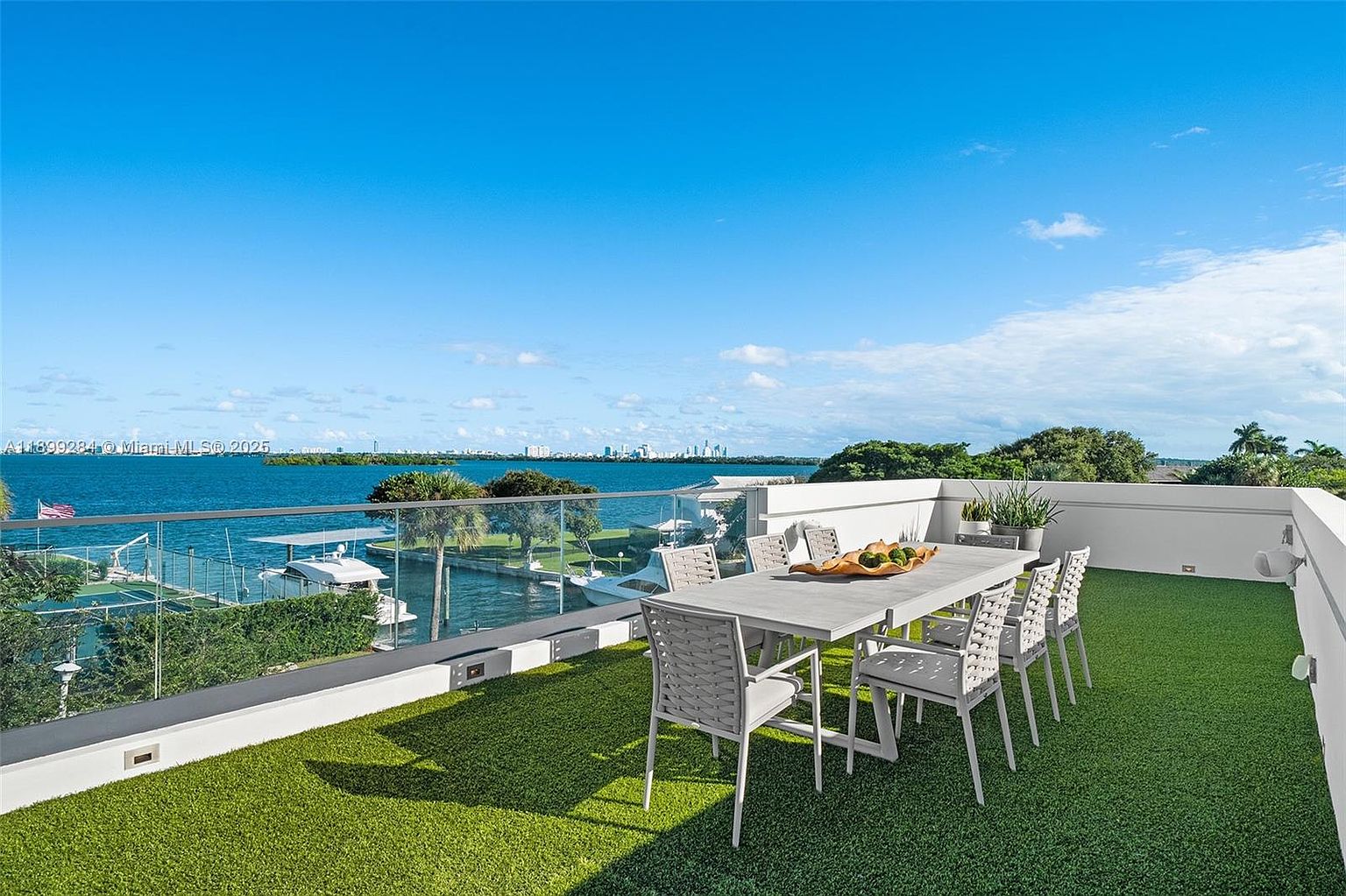 This image showcases a luxurious rooftop patio/deck with artificial grass, a modern dining set, and a stunning waterfront view. The clear glass railing provides an unobstructed panorama of the bay, with boats and the city skyline visible in the distance. The overall impression is one of upscale outdoor living and relaxation.