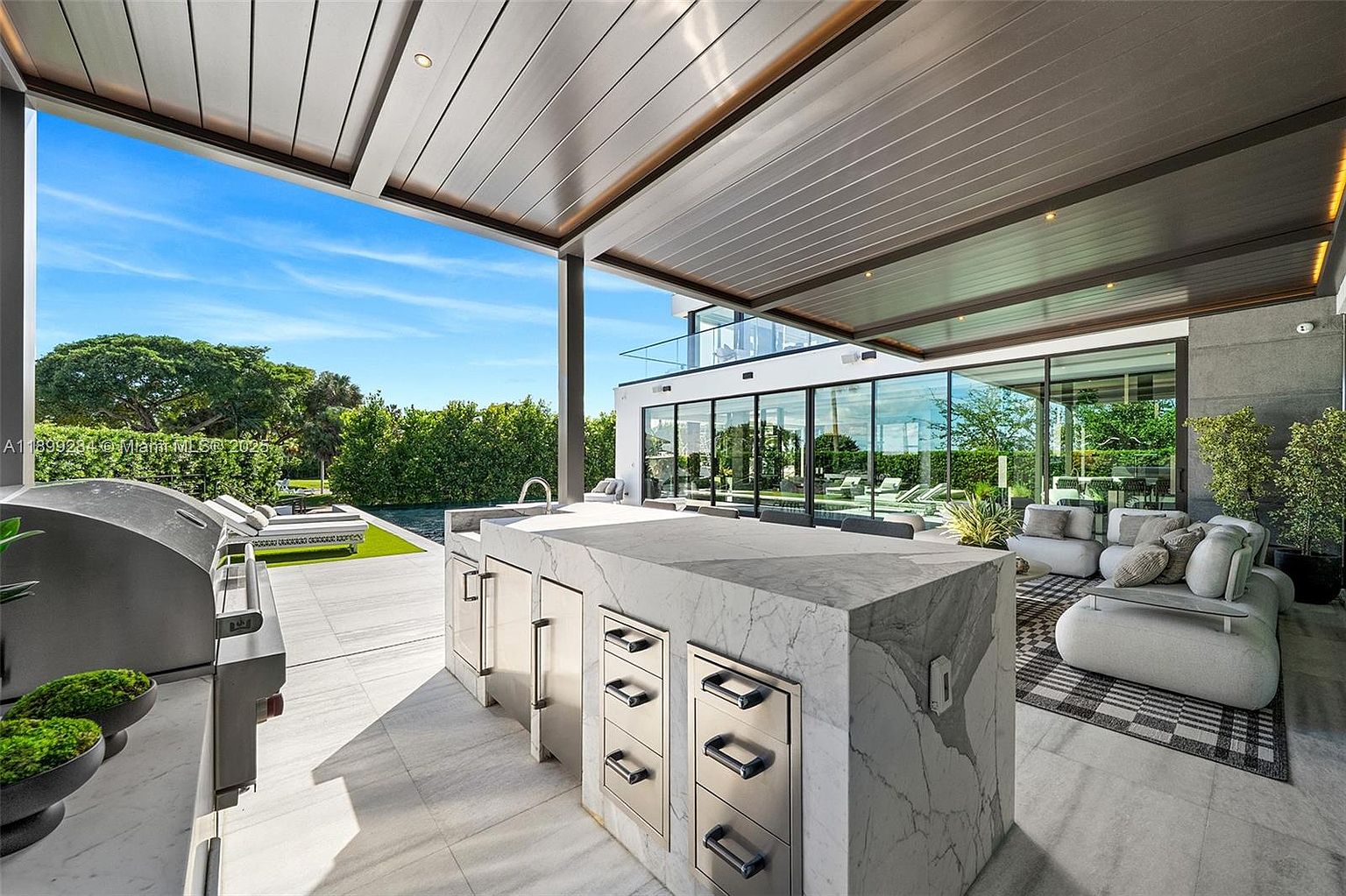 This image showcases a luxurious outdoor patio area, featuring a modern outdoor kitchen with a marble island and stainless steel appliances. A comfortable seating area with plush couches and a stylish rug is visible, while the backdrop includes a sleek, modern home with large glass windows overlooking a pool and manicured lawn. The overall impression is one of sophisticated outdoor living and entertaining.