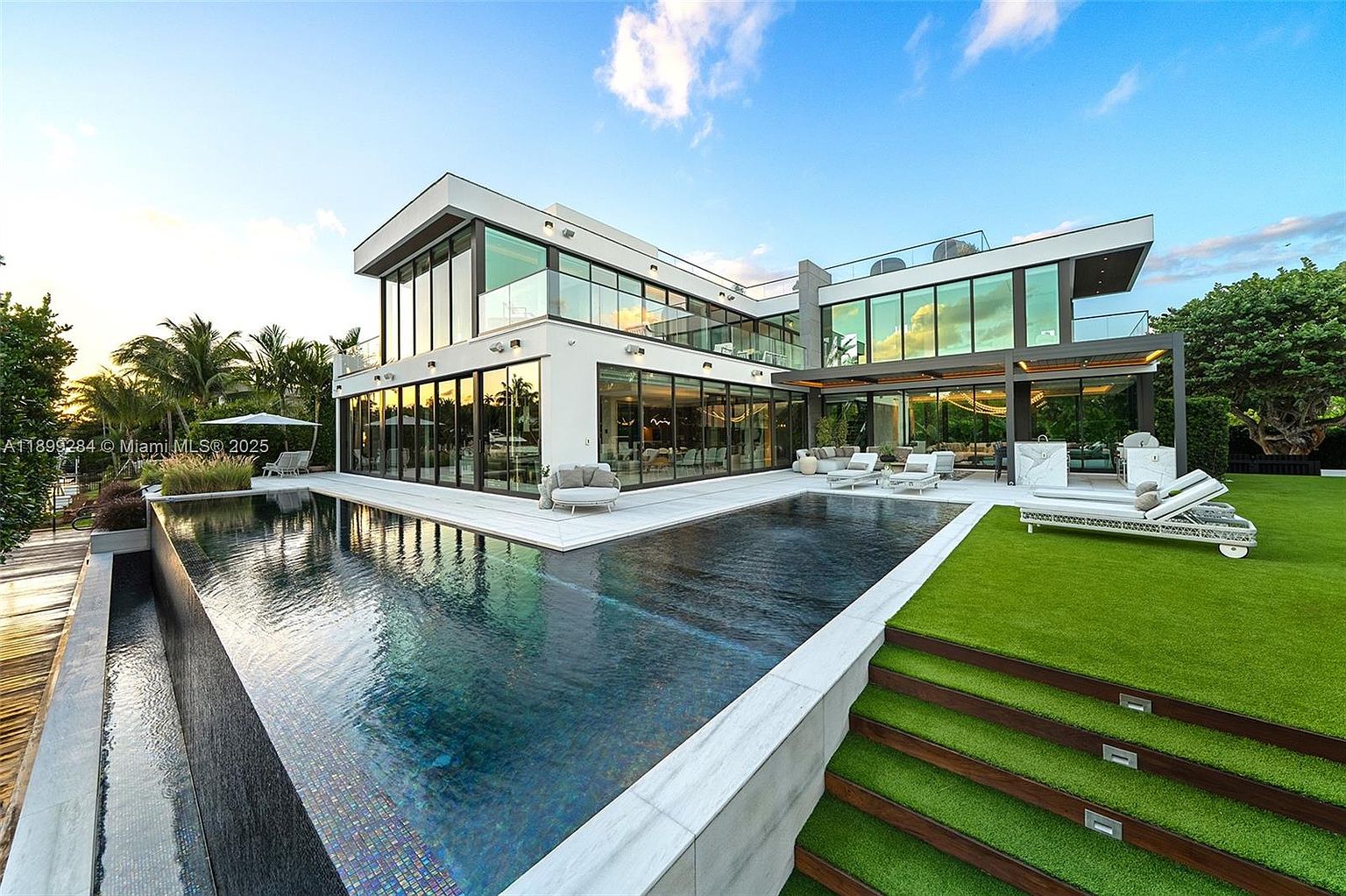 This image showcases the rear exterior of a modern luxury home, emphasizing the stunning pool and outdoor living space. The pool features a dark, reflective surface and is bordered by a sleek, white patio. Adjacent to the pool is a manicured lawn with steps leading up, creating an inviting outdoor oasis perfect for relaxation and entertainment.