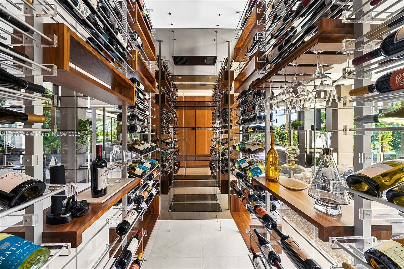 This is an interior shot of a modern wine cellar, showcasing a symmetrical design with wooden shelves and metal supports. Bottles of wine are neatly arranged on both sides, with glass racks for wine glasses and decanters adding to the sophisticated ambiance. The perspective is a straight-on view, emphasizing the depth and organization of the cellar.