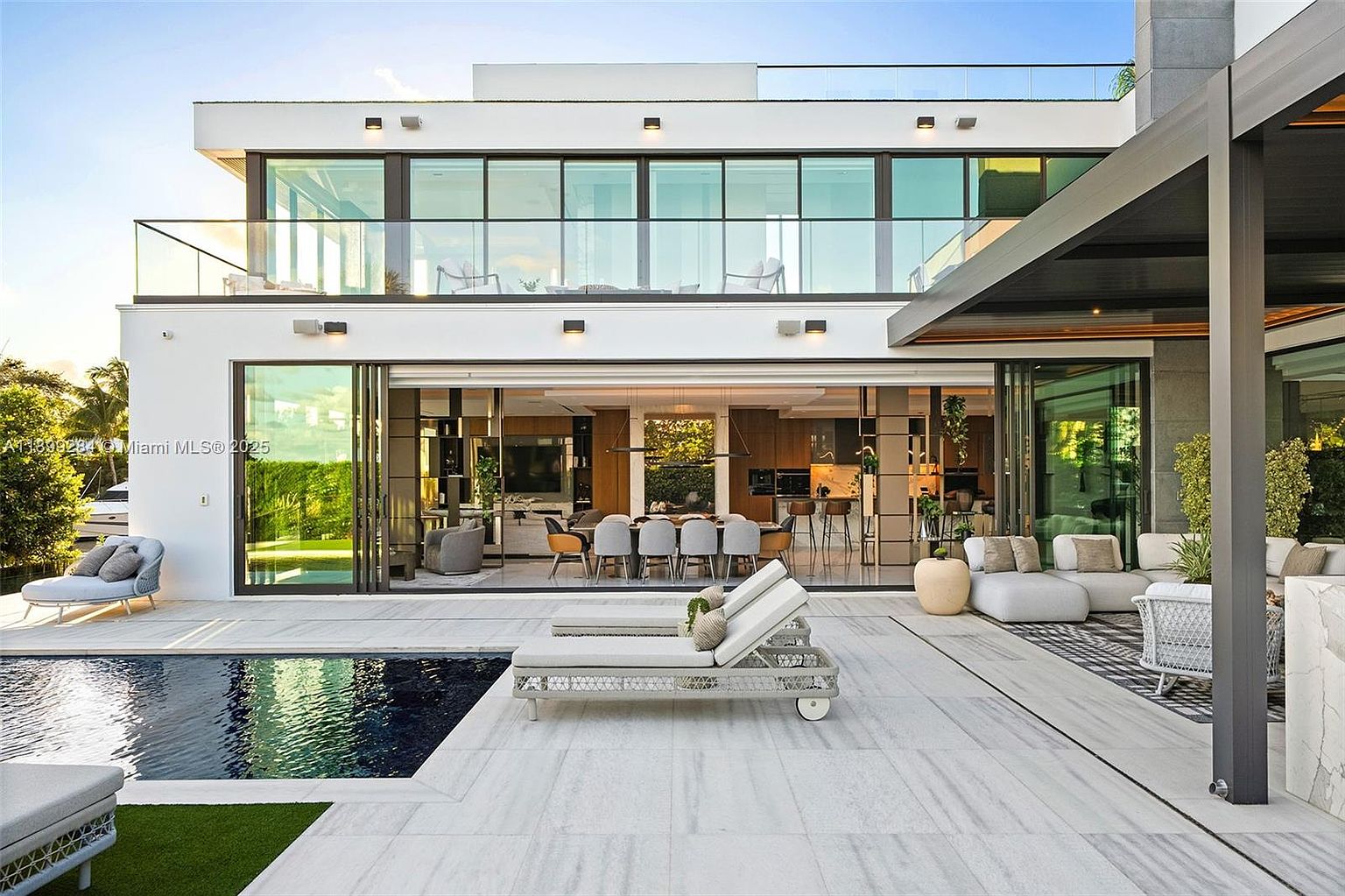 This image showcases the rear exterior of a modern luxury home, featuring expansive glass sliding doors that open to a combined living and dining area. A sleek pool and patio area with lounge chairs provide an inviting outdoor space, while the upper level boasts a balcony with glass railings. The overall design emphasizes indoor-outdoor living and contemporary elegance.